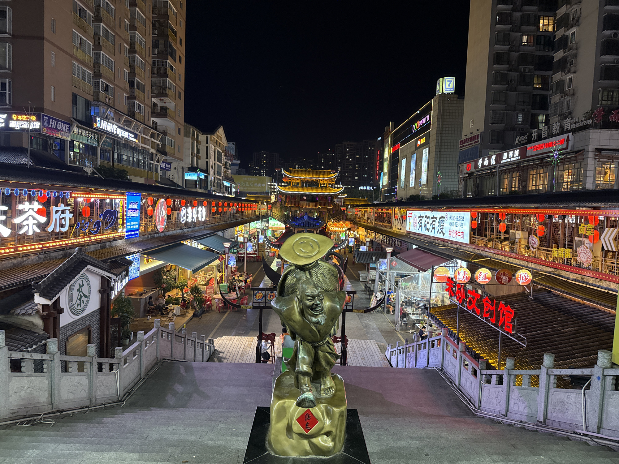 This night scene captures a vibrant, multi-level commercial street or market in Zhangjiajie, China, under a dark, clear night sky. The area is brightly illuminated by numerous artificial lights, neon signs, and traditional red lanterns, creating a lively and festive atmosphere.
In the immediate foreground, a prominent bronze-colored statue of a broadly grinning man stands at the top of a wide stone staircase. He wears a large hat and appears to be carrying something substantial on his back. A red square with a gold character, likely "福" (Fú, meaning fortune or good luck), is visible on the statue's base.
Flanking the staircase and extending into the midground are elevated walkways lined with various shops and restaurants. Many of these establishments feature traditional Chinese tiled roofs and intricate architectural details, contrasting with the modern high-rise buildings visible in the background. Numerous people are present, seen dining at outdoor tables, socializing, and walking along the market pathways, suggesting a bustling evening activity.
Visible text on the left includes "HIONE 乡莎瓷砖" (HIONE Xiangsha Tiles), "三茶府" (San Cha Fu - Three Tea House/Mansion), "海外有" (Overseas/Foreigners Have), and "热卖中" (Hot Sale/Selling Well). On the right side, a "7" logo (likely 7-Eleven), "开智教育" (KaiZhi Education), "有肥有瘦" (You Fei You Shou - Fat and Lean, possibly for a food stall), "鲜切烤牛肉" (Fresh Cut Roast Beef), and "榕树文创馆" (Rongshu Wenchuan Guan - Banyan Tree Cultural & Creative Hall) are visible, indicating a diverse range of businesses. The overall scene conveys a sense of cultural richness and urban dynamism.