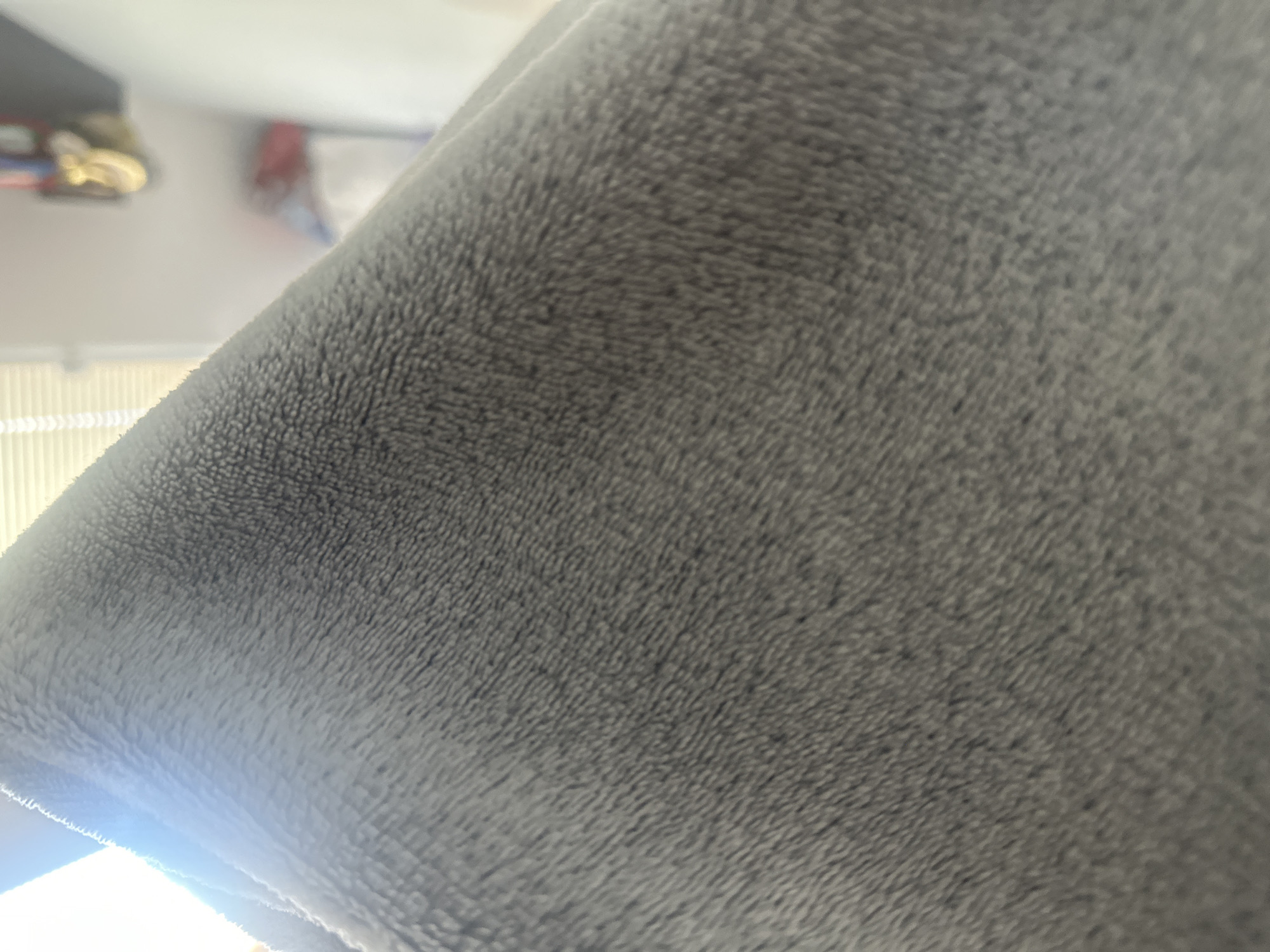The image is a close-up of a grey, plush blanket or fabric, with a bright light source reflecting off the surface on the left side. The texture of the material is visible, showing a soft, fuzzy pile. In the background, out of focus, there is a hint of a room with blinds on a window and some objects on a shelf. No specific location within the city of Shumen, Bulgaria, can be identified from this image.