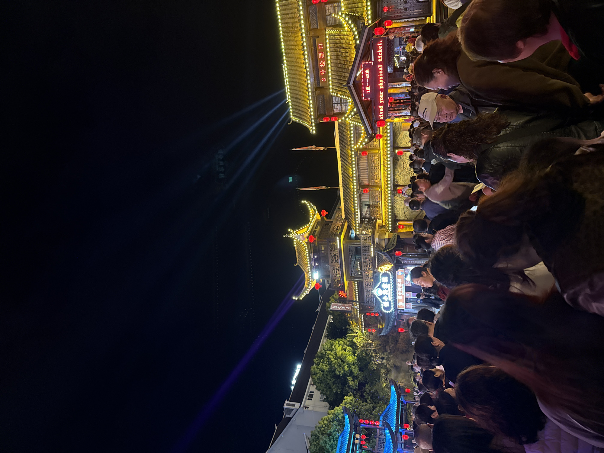 This media file captures a bustling night scene in Wulingyuan, China, featuring traditional Chinese architecture illuminated by numerous lights and a large crowd of people.
In the foreground, a dense gathering of people fills the lower portion of the image, mostly viewed from behind or from above their heads. They appear to be walking or waiting in line, suggesting a popular attraction or event. The crowd is casually dressed, and while individual faces are mostly obscured, the sheer number indicates a lively atmosphere.
The mid-ground and background are dominated by ornate, multi-tiered traditional Chinese buildings. These structures are extensively decorated with warm yellow string lights outlining their roofs and eaves, and numerous red lanterns hanging from their facades. From behind one of the main buildings, several bright spotlights project beams upwards into the dark night sky. Some green foliage is visible, also illuminated by the surrounding lights. Further back, another structure features blue decorative lights.
The time of day is clearly night, characterized by the dark sky and the vibrant artificial lighting creating a festive ambiance. The setting evokes a cultural or historical district, likely an entertainment or performance venue given the signs.
Visible text includes a red LED sign on one of the main buildings displaying Chinese characters "2 号检票口" (No. 2 Ticket Gate/Check-in Counter) and "1楼—45座" (1st Floor - 45 seats), indicating a ticketing or entry point for an event or show. Partially visible English text on the same sign reads "received your physical ticket," suggesting instructions for attendees.