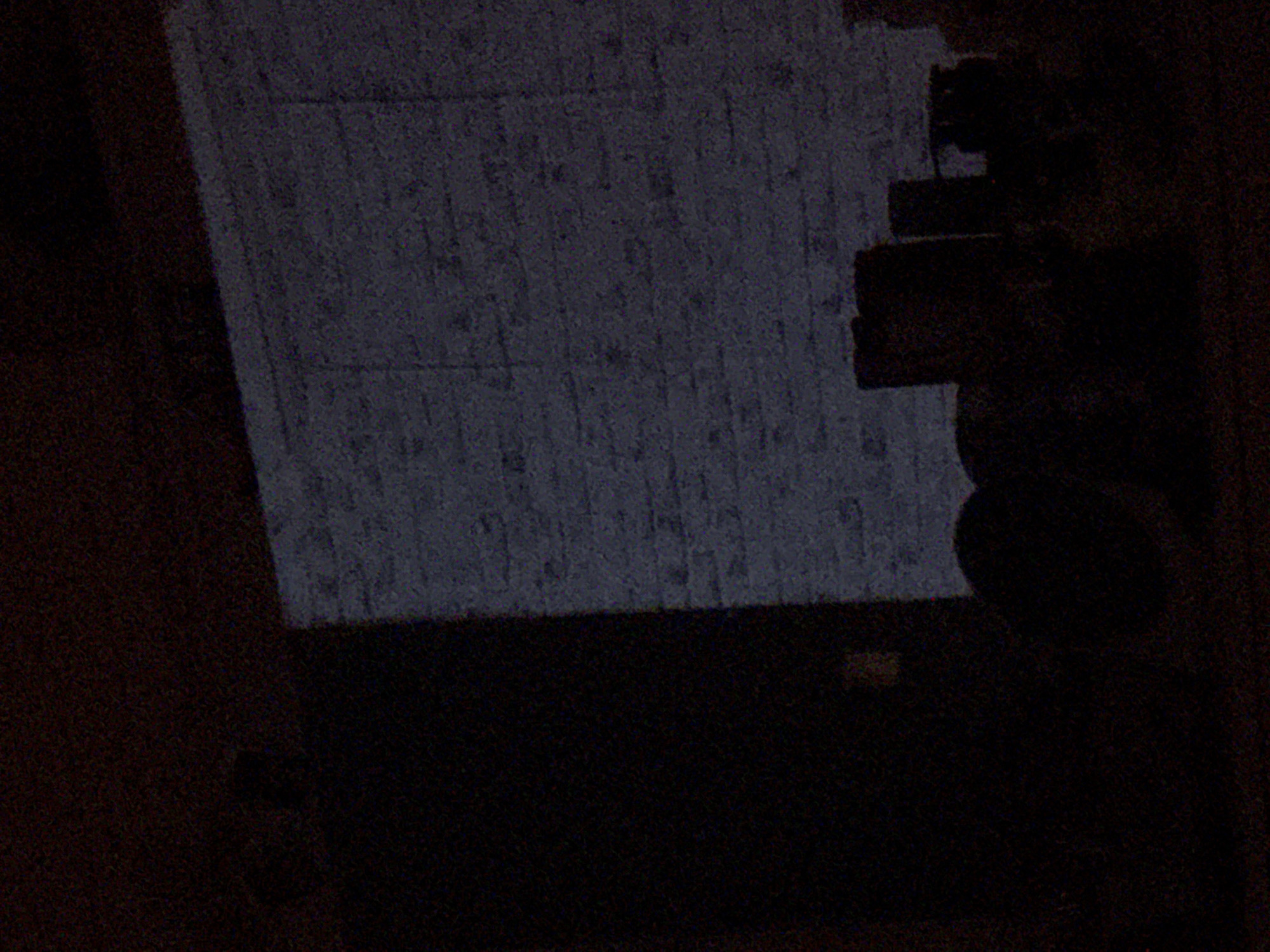 The extremely dark and grainy image, likely taken in low light conditions, depicts an indoor scene in Sofia, Bulgaria. The central focus is a large, rectangular surface, possibly a screen or a wall acting as a projection surface, which emits a faint, cool blue-grey light. This surface occupies the upper left and central portions of the frame. The details on this screen are highly indistinct due to the low resolution and heavy noise, appearing as a mottled, textured pattern with some vertical striations, possibly representing text or graphics that cannot be read.
To the immediate right of the illuminated screen, a dark, shadowy, and somewhat blocky object or piece of equipment is visible. Its exact nature is unclear, but it could be a projector, a camera, or part of a mounting system, silhouetted against the ambient darkness. The rest of the image, particularly the bottom and far right, is almost entirely enveloped in deep shadows, rendering any other features indiscernible. The overall setting suggests a room prepared for a presentation, a movie viewing, or some form of display, conducted in near-total darkness, indicative of a night-time event or a controlled environment. No discernible text or people are visible.