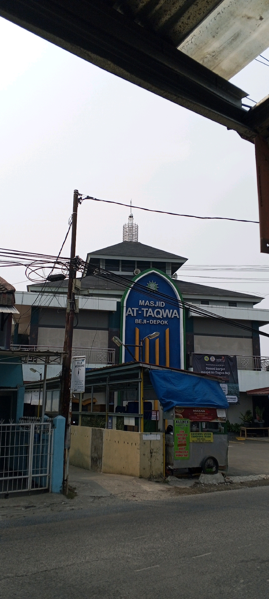 The image shows a street view in Depok, Indonesia, featuring the "Masjid At-Taqwa Beji-Depok" (At-Taqwa Mosque Beji-Depok). The mosque has a dark roof and a distinctive blue facade with the mosque's name in white lettering. A tall, slender tower with a coiled structure tops the mosque. In the foreground, there are utility poles with a dense network of electrical wires crisscrossing the scene. To the right of the mosque entrance, a food cart is visible with blue tarpaulin covering. The cart displays various food items and prices in Indonesian. A portion of the street and a sidewalk with a light blue fence are also present. The sky is overcast.