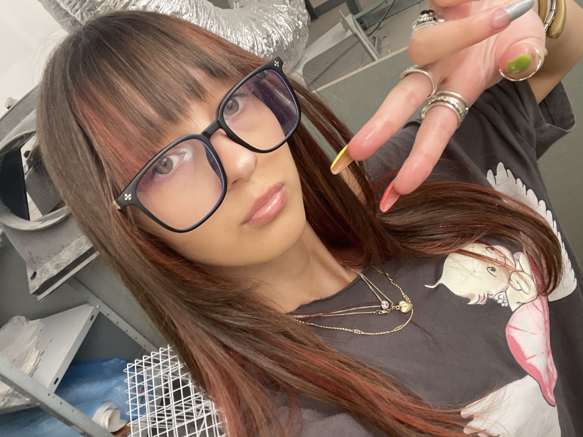 A young woman with long, reddish-brown hair and bangs is taking a selfie in what appears to be a workshop or storage area. She is wearing glasses with black frames and is showing a peace sign with her right hand. Her fingernails are long and decorated with various colors and designs, including pink, yellow, green, and gray. She is also wearing several rings on her fingers and a necklace with a heart pendant. She is wearing a dark t-shirt with a cartoon image of a baby on it. The background shows shelves, various metal objects, and what looks like insulation or foil. The image does not provide any specific clues to suggest the location is Raanana, Israel.