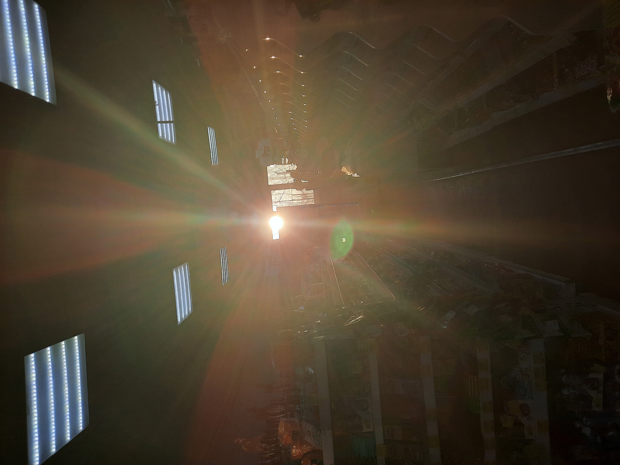 This rotated image, likely captured in Krasnogorskoye, Russia, presents the interior of what appears to be a long hall, corridor, or gallery. The perspective is angled upwards or along the length of the space, but the photograph itself is rotated 90 degrees clockwise, making elements that would naturally be vertical (like walls) appear horizontal.
The scene is dominated by a powerful, central light source, which could be an overhead light fixture, a skylight, or the sun entering from a window. This intense light creates a dramatic, star-shaped lens flare with prominent radial streaks of green, orange, and red, as well as a distinct circular green artifact. The flare significantly obscures details in the center and lower-right parts of the image, while the surrounding areas remain relatively dark.
On what appear to be the side walls (visible at the top and bottom of the rotated image), several rectangular LED light panels are mounted. These fixtures feature multiple vertical strips of bright white LEDs. Closer panels are sharply defined, while those receding into the distance appear smaller and less distinct. Above, a structured or possibly corrugated ceiling stretches into the distance, reflecting light and casting faint patterns. Further into the background, beneath the ceiling structure, vague and dark shapes suggest shelves or displays laden with numerous items, possibly posters, stickers, or merchandise, hinting at a commercial or exhibition setting. No people or legible text are visible. The image conveys a dramatic atmosphere due to the stark contrast between the blinding light and the deep shadows.