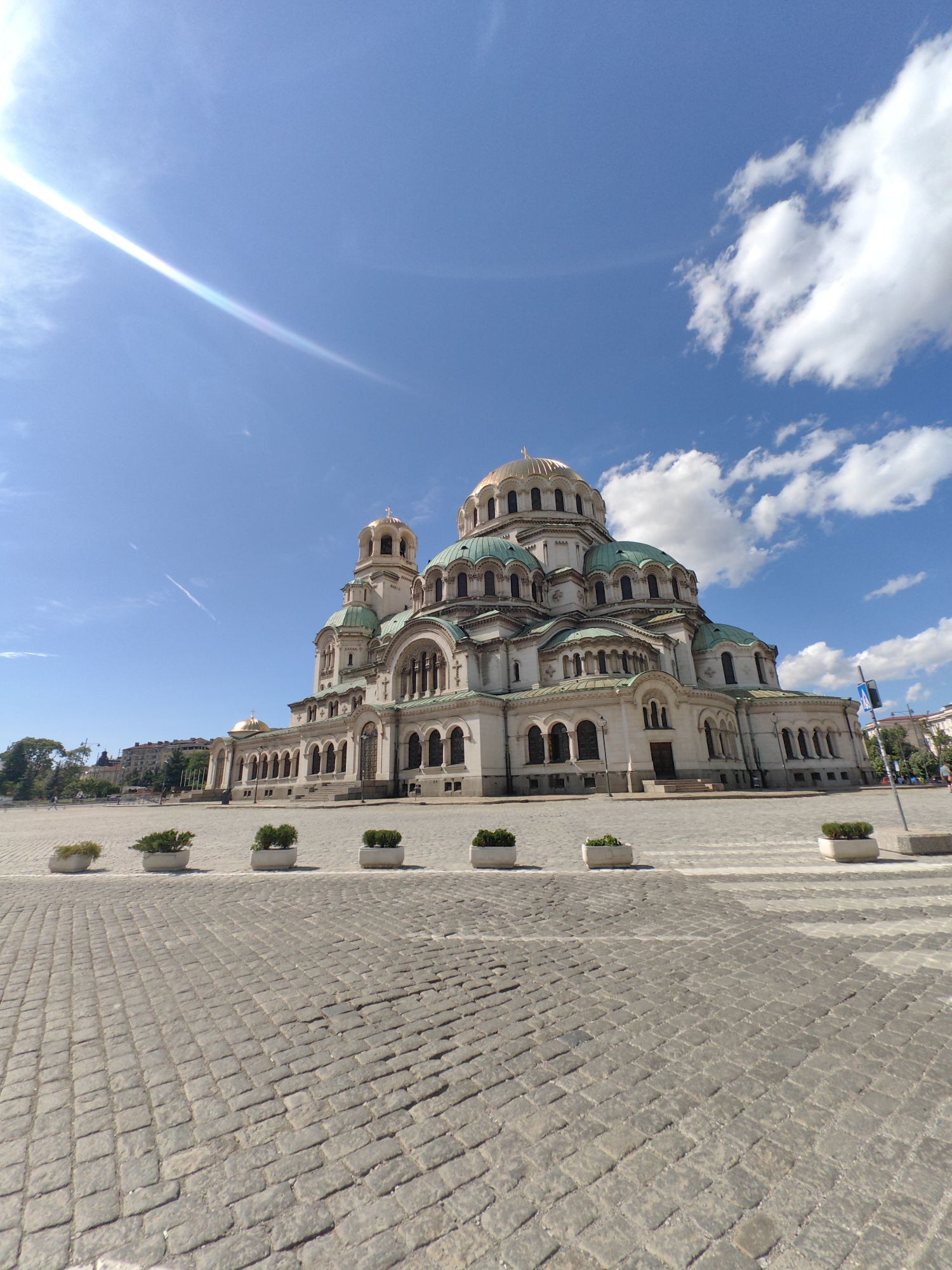 A wide-angle, low-angle shot captures the monumental Alexander Nevsky Cathedral in Sofia, Bulgaria, under a bright blue sky dotted with fluffy white clouds. The sun, positioned in the upper left quadrant, casts a lens flare across the sky. The cathedral, a prominent landmark, features a large central golden dome and several smaller green domes, all adorned with crosses. Its facade is characterized by numerous arched windows and intricate architectural details. In the foreground, a cobblestone plaza stretches out, with rows of white planters containing green shrubs spaced evenly across its surface. The plaza is largely empty, with only a few distant figures visible, suggesting a quiet, perhaps early morning or late afternoon, scene. The overall atmosphere is one of grandeur and serenity.