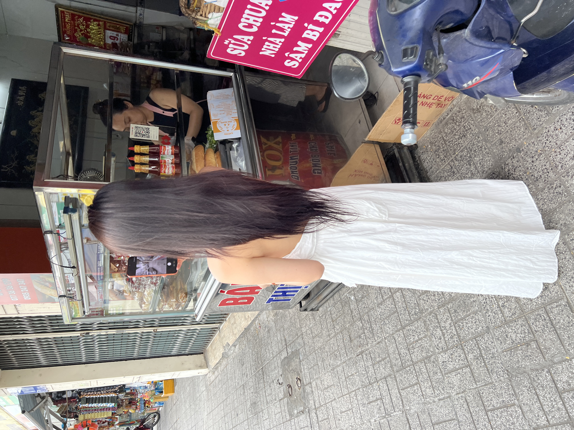 The image shows a street-level view of a small shop in Nha Trang, Vietnam, likely a bakery or food stall. The main subject is a young woman with long dark hair, wearing a white dress, who is standing with her back to the camera. She is holding up a smartphone, seemingly taking a photo of the shop's display.
The shop itself is characterized by a glass display case filled with various items, including bread loaves, bottles of sauce, and packaged goods. Behind the counter, a woman with her hair tied up is visible, wearing an apron and gloves, and appears to be serving a customer or preparing food. A blue scooter is parked to the right of the shop, with a sign attached to it.
Prominent signage is visible in Vietnamese. A pink sign above the shop reads "SỮA CHUA NHÀ LÀM SÂM BÍ ĐA," which translates to "Homemade Yogurt, Bitter Melon Juice." Another sign near the scooter displays "SẴN LÀNG NGHE TAY" and "CÀNG DỄ VỚ," suggesting readiness to serve or handle orders. There are also numbers on a red sign and other indistinct text on various signs and packaging within the display case.
The setting appears to be daytime, with natural light illuminating the scene. The ground is paved with textured tiles. The overall impression is a bustling street scene in Vietnam, with a focus on the daily operations of a local food business and a customer engaged with their phone.
