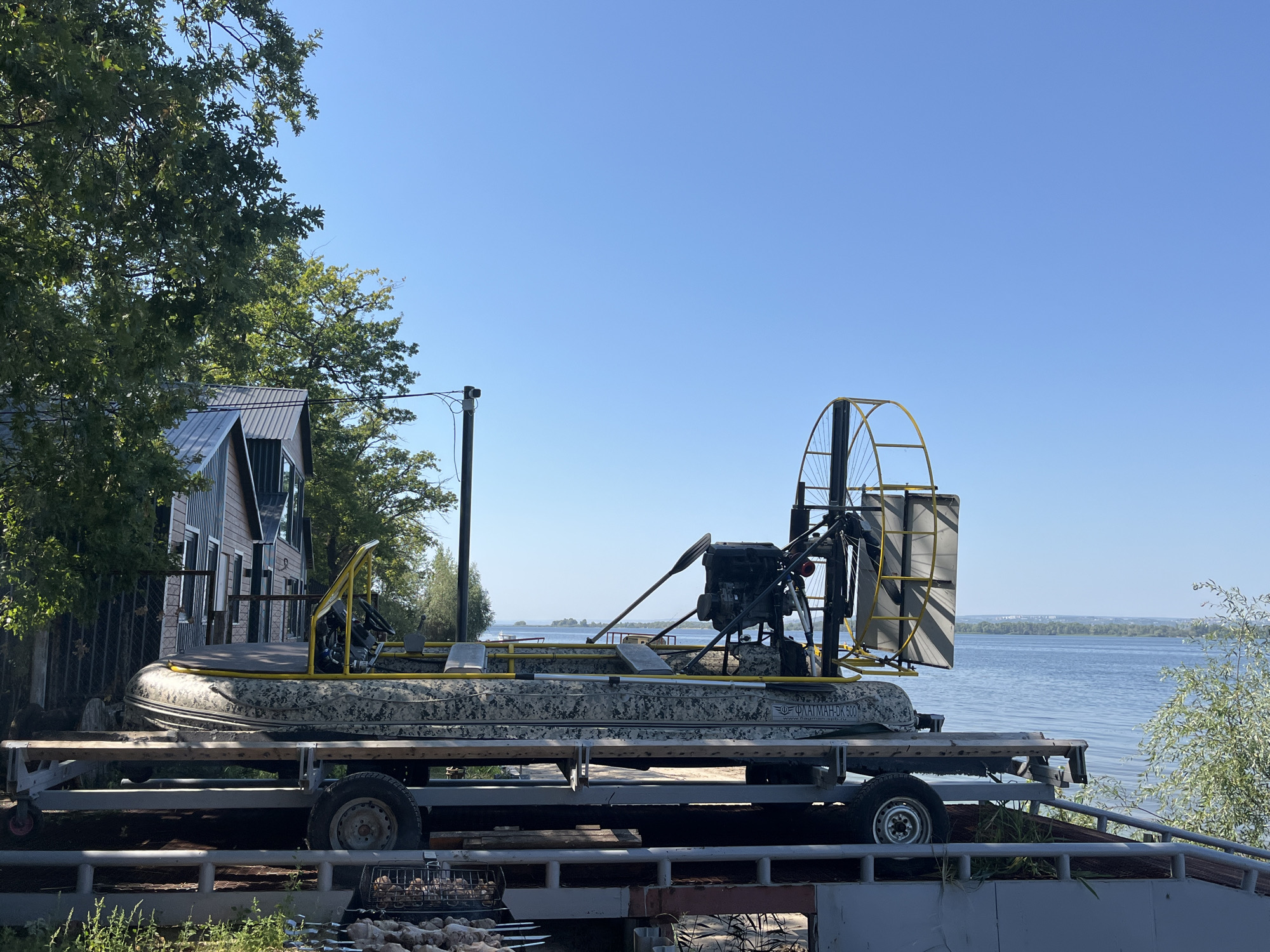 An airboat with a camouflage pattern sits on a trailer. The airboat has a yellow frame around the controls and a large fan at the back. The trailer is parked next to a building with a metal roof and wood siding, likely in Shumeika, Russia, as indicated by the text on the boat which reads "ФЛАТМАН-ДК 500", a Russian brand. A body of water, possibly a river or lake, is visible in the background, with trees lining the shore and a clear blue sky overhead.