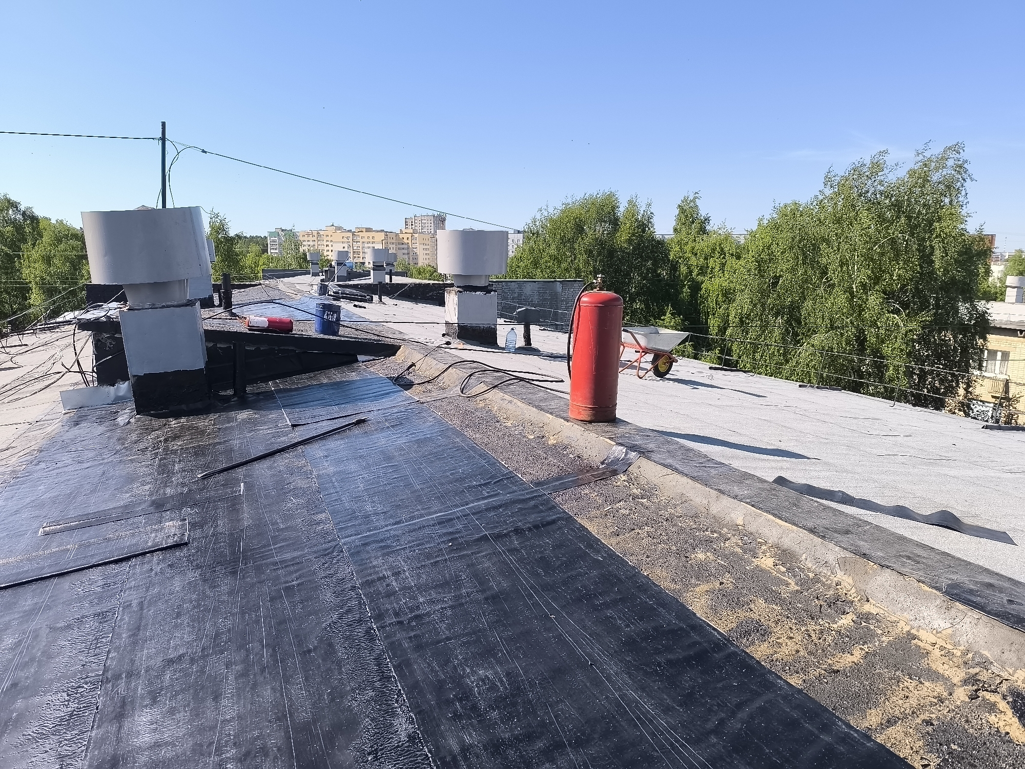 The image shows a flat rooftop in an urban area. The roof is covered with a mix of dark, presumably newly installed, rolled roofing material and older, light gray roofing. Several ventilation stacks with cylindrical tops are visible, along with a red fire extinguisher and a wheelbarrow. In the background are trees and multi-story buildings under a clear blue sky. A power line or cable stretches across the upper part of the image. The location is Unknown, Unknown.