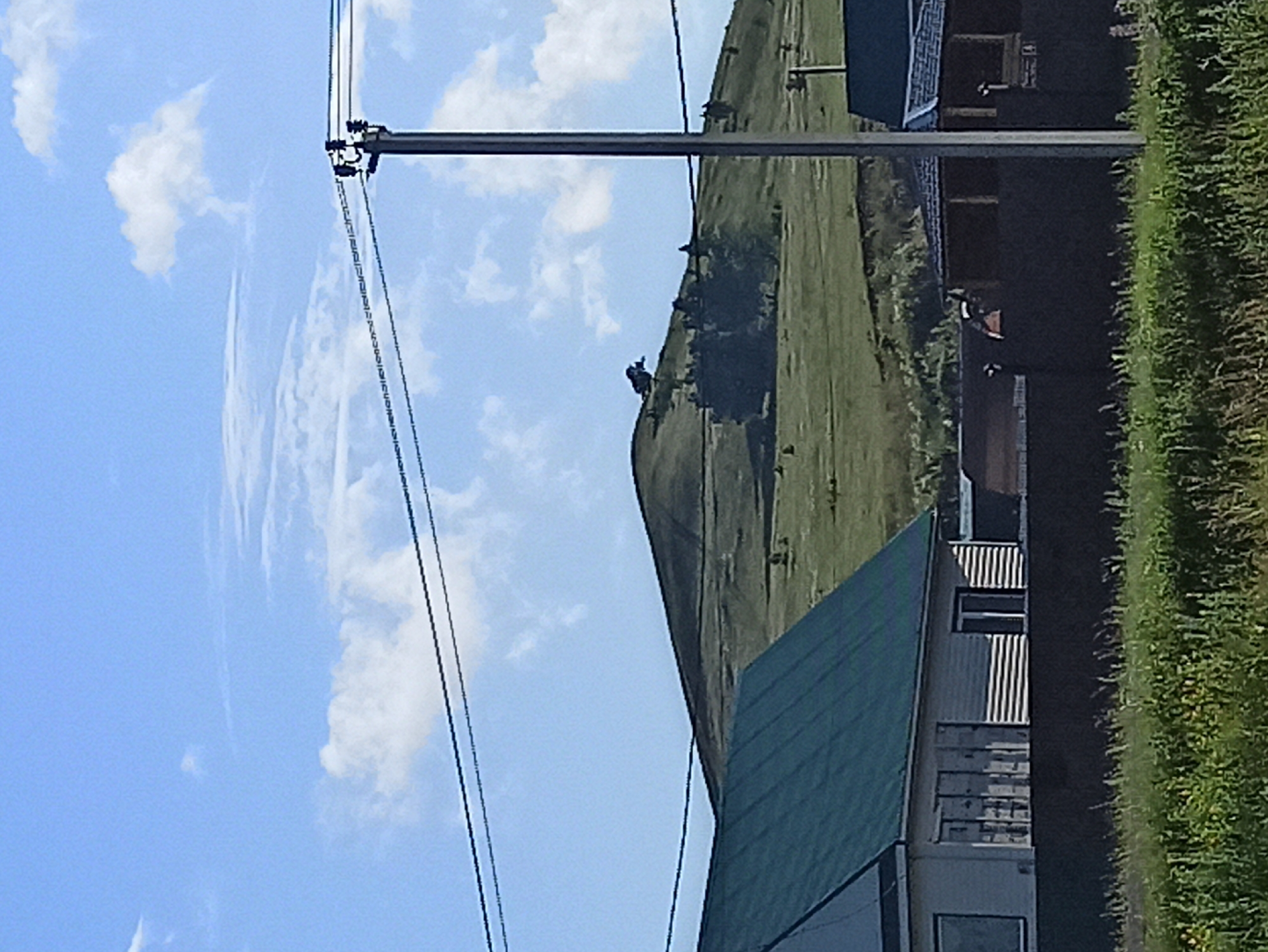 This media file shows a rural scene in Isyanovo, Russia. The image is oriented vertically. In the foreground, a modern building with a dark green roof is visible, alongside a grassy verge. Further back, a steep, grassy hill dominates the landscape, with a few scattered trees or bushes. A power line runs horizontally across the upper portion of the image, with poles and wires clearly discernible. In the distance, a few buildings with dark roofs and windows are nestled against the hillside. The sky is blue with scattered white clouds, suggesting it is daytime and the weather is fair. The overall impression is one of a quiet, perhaps remote, rural setting. No people are visible in the frame. There is no visible text in the image.
