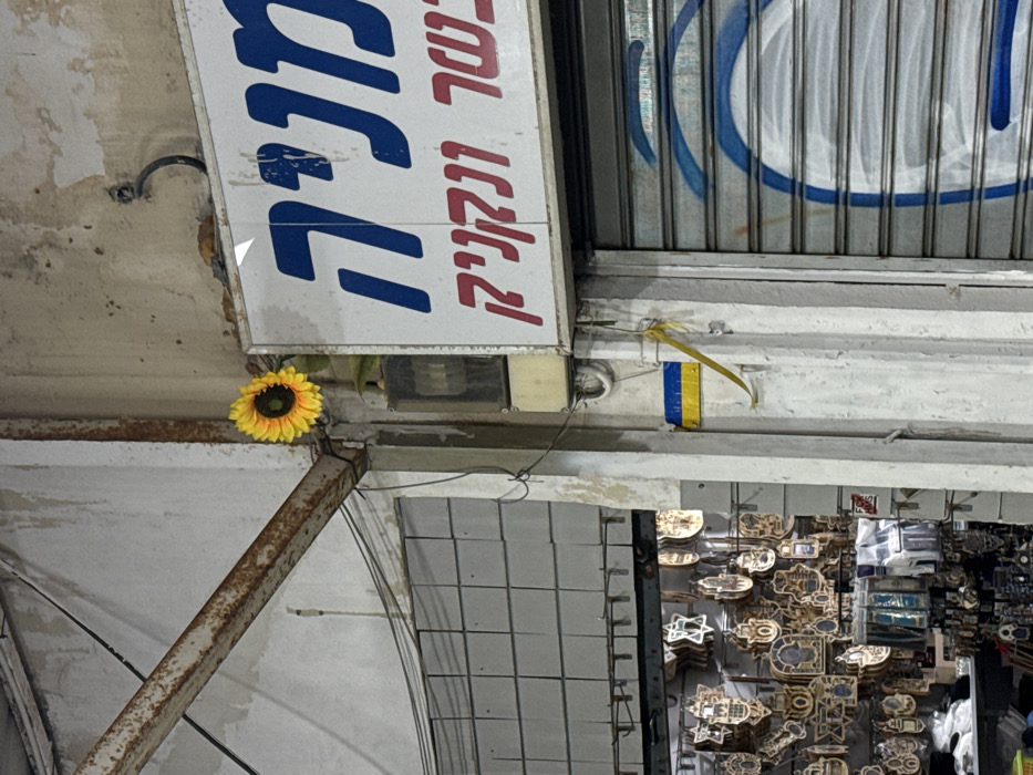 The image displays a close-up, static urban scene in a commercial area of Tel-Aviv, Israel. The setting features aged building facades with utilitarian elements.
On the right, a shopfront is closed with a corrugated metal rolling shutter, which exhibits blue graffiti in circular patterns. Above the shutter, a rectangular white sign with Hebrew text is affixed. The upper blue text reads "מניה" (Manya) and "בשר" (Basar - meat), while red text below states "ונקניק" (Ve'Neknik - and sausage), indicating a butcher shop or deli. An artificial yellow sunflower with a dark brown center is positioned above the sign, attached to the wall or a fixture. Below the sign and to its left, a small electrical box is visible.
A structural white column, showing signs of wear and dirt, separates the right shop from an adjacent establishment on the left. On this column, a small rectangular sticker or sign depicting the blue and yellow colors of the Ukrainian flag is affixed, with a thin yellow ribbon or string hanging from it.
To the left of the column, a shop entrance is open, revealing merchandise. Inside, numerous wooden and possibly metal decorative items, including shapes like Stars of David and Hamsas, are hanging on display. Additional packaged items are visible on lower shelves.
Structural elements include a rusted metal I-beam or support beam crossing the upper left of the frame, and a tiled section on the upper wall of the left-side building. Electrical conduits and wires are visible running along the walls and beam. No people or active interactions are present in the scene.