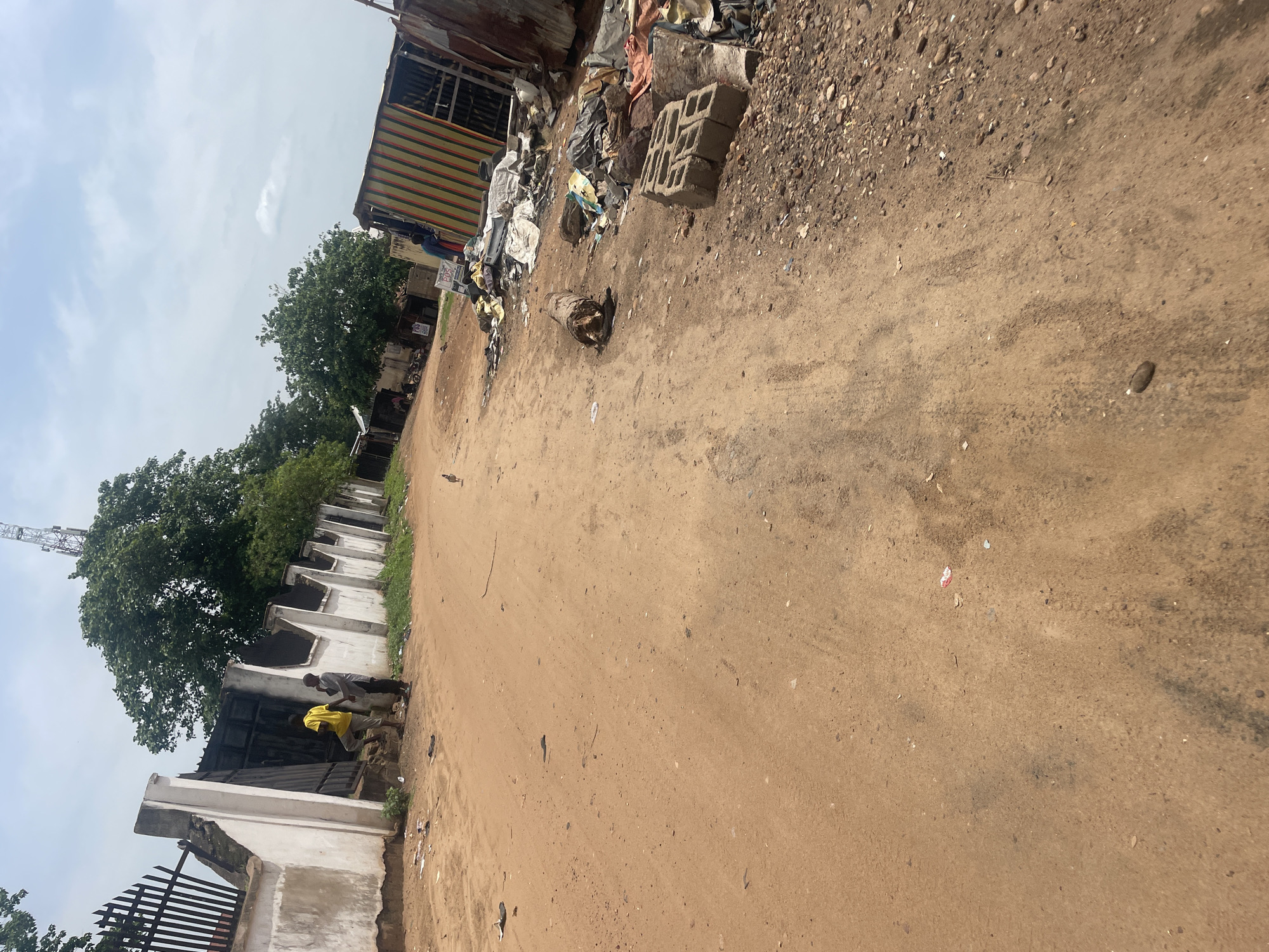 The image depicts a dusty, unpaved street in Lokoja, Nigeria. In the foreground, the sandy road dominates, with scattered debris including small rocks, twigs, and some small pieces of trash. In the mid-ground to the left, two young men are standing near a white-washed wall with a black metal gate. One is wearing a yellow t-shirt and shorts, and the other is wearing a grey t-shirt and dark trousers. A small bird is visible on the road in the mid-ground. To the right, in the background, is a pile of discarded materials including plastic sheeting, what appears to be a metal shipping container painted with colorful vertical stripes, and other refuse. Several cinder blocks are also present near the debris. The environment is characterized by a dry, arid appearance with sparse vegetation and a cloudy sky overhead. The scene appears to be on the outskirts of a residential or commercial area, possibly a market or informal settlement, given the presence of makeshift structures and accumulated waste. A telecommunications tower is faintly visible in the far distance above the trees.