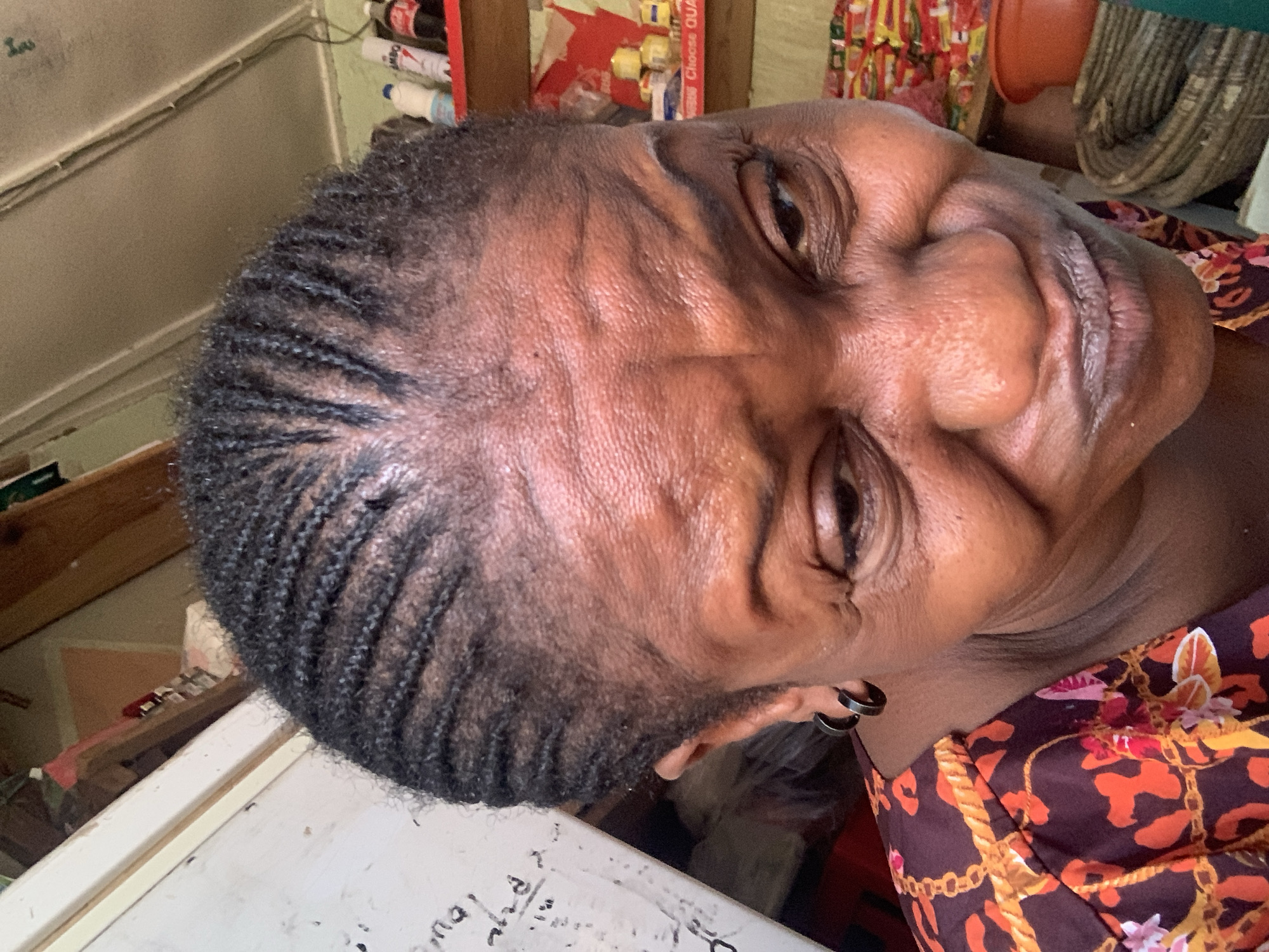 A medium shot captures an adult African woman with cornrow braids, viewed from slightly above at a tilted angle. She is wearing a patterned brown and orange garment. The background suggests an indoor environment, possibly a home or shop. Details like white boards, shelves with various items, and tubing are discernible. The location is suggested to be Sabon Tasha, Nigeria, although no specific landmarks or identifying details confirm the location within the scene.
