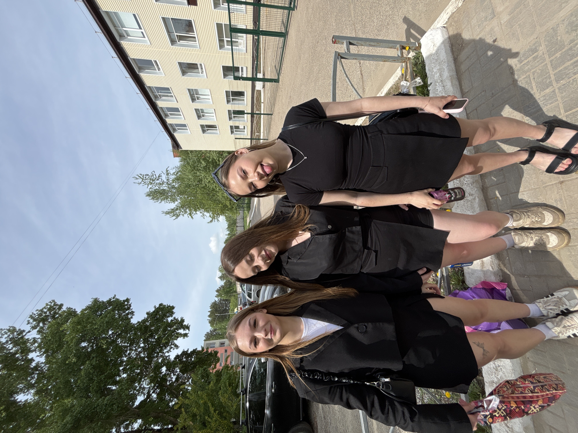 Three young women stand side-by-side outdoors. The woman on the right wears a black dress, black sandals, and sunglasses on her head. She holds a phone in one hand and a beverage container in the other. The woman in the center wears a black outfit and off-white sneakers. The woman on the left wears a white shirt under a black blazer and white socks with sneakers. A black car is parked behind her, and she holds a purse. A bag and other items are on the ground near the center woman's feet. The background shows a building with rectangular windows, green fencing, trees, and a paved area. The location is in the city of Chaikovsky, Russia.