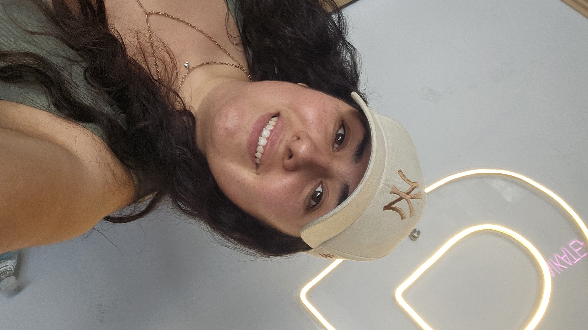 A young woman with long, dark, wavy hair smiles warmly at the camera in what appears to be an indoor setting. Her head is tilted, giving the image a casual, selfie-like feel. She is wearing a light-colored (possibly beige or off-white) baseball cap with a prominent, dark-colored embroidered "NY" logo, indicative of the New York Yankees. A delicate gold chain necklace with a small, clear gemstone or charm is visible around her neck and across her chest. She is also wearing a light-colored top, possibly olive or green.
The background is a plain, light-colored wall (white or light gray). A bright neon light sign is visible in the lower right portion of the image. The sign forms a curved shape, and part of it glows with a warm yellow/orange light, while another section, which spells out visible text, glows pink/purple. When oriented correctly, the glowing text appears to read "IKALE" or "KALE", likely part of a name or brand. A small water bottle with a white cap is subtly visible in the bottom left corner. The overall ambiance suggests a modern, relaxed environment, possibly in Puebla, Mexico.