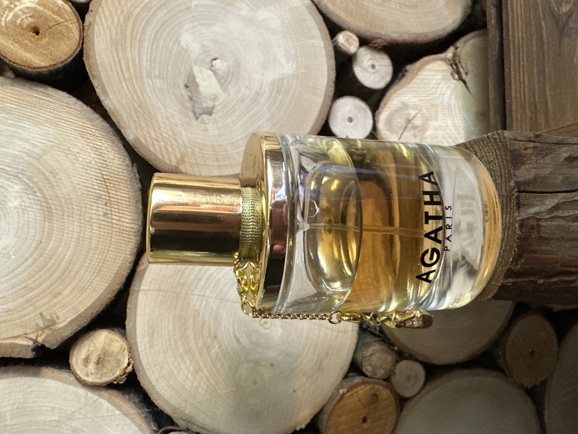 A perfume bottle with a gold cap and chain detail sits on a wooden surface in front of a backdrop of stacked wood slices. The perfume bottle is made of clear glass and contains a golden liquid. The brand name "AGATHA PARIS" is printed in black lettering on the bottle. The wood slices in the background display natural grain patterns and vary in size. The overall scene is rustic and natural. The provided information about the location in Norviliškės, Lithuania is not visually supported by the image content.