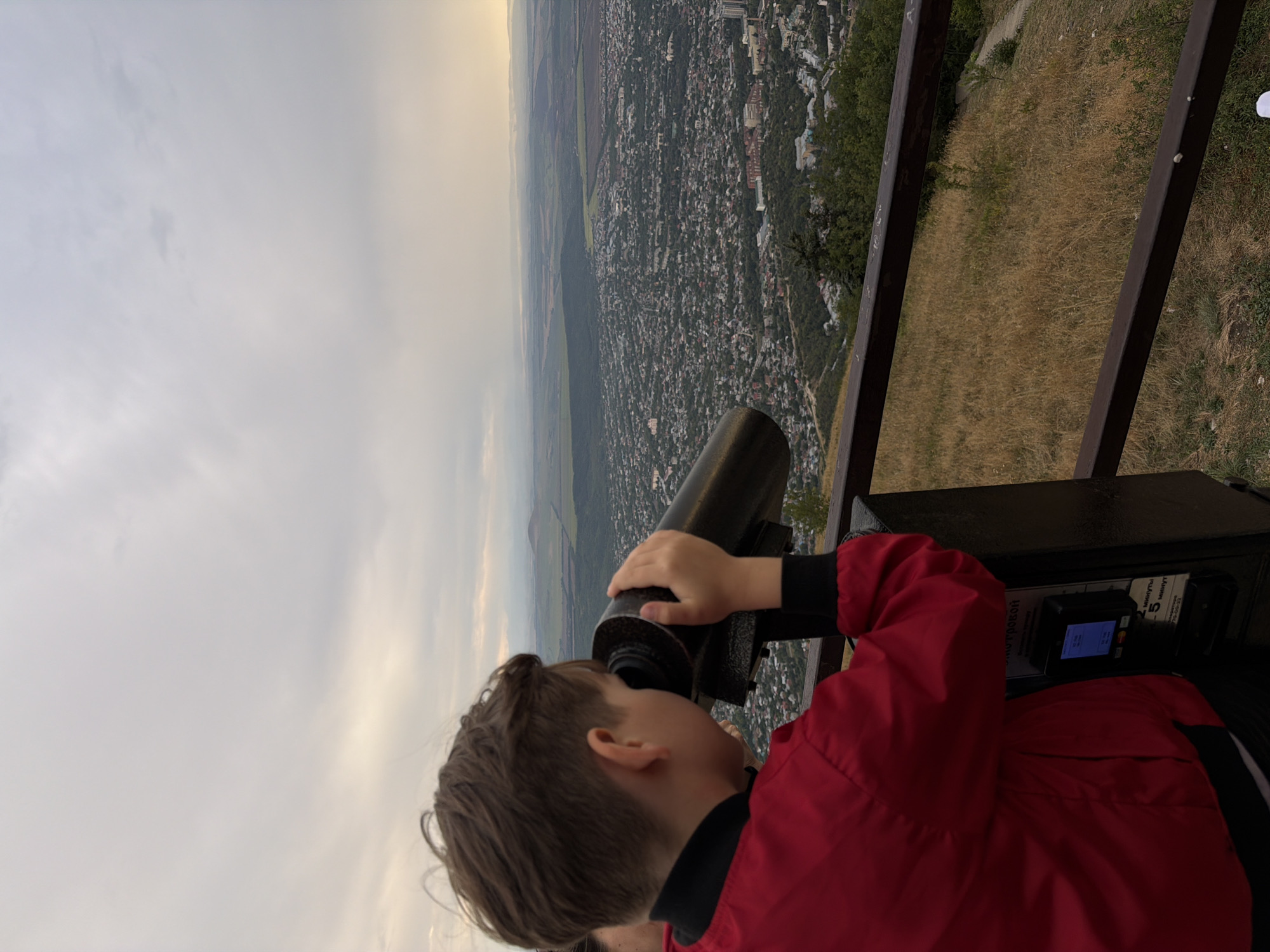 A young boy wearing a red jacket is looking through a coin-operated telescope at a panoramic view of the city of Goryachevodsky, Russia, from a hilltop. The sky is overcast with the sun setting, casting a warm glow on the distant landscape. The telescope is mounted on a dark stand with a digital display and coin slot. A metal railing is in the foreground, and dry grass covers the hillside. The city below is densely populated with buildings and trees.