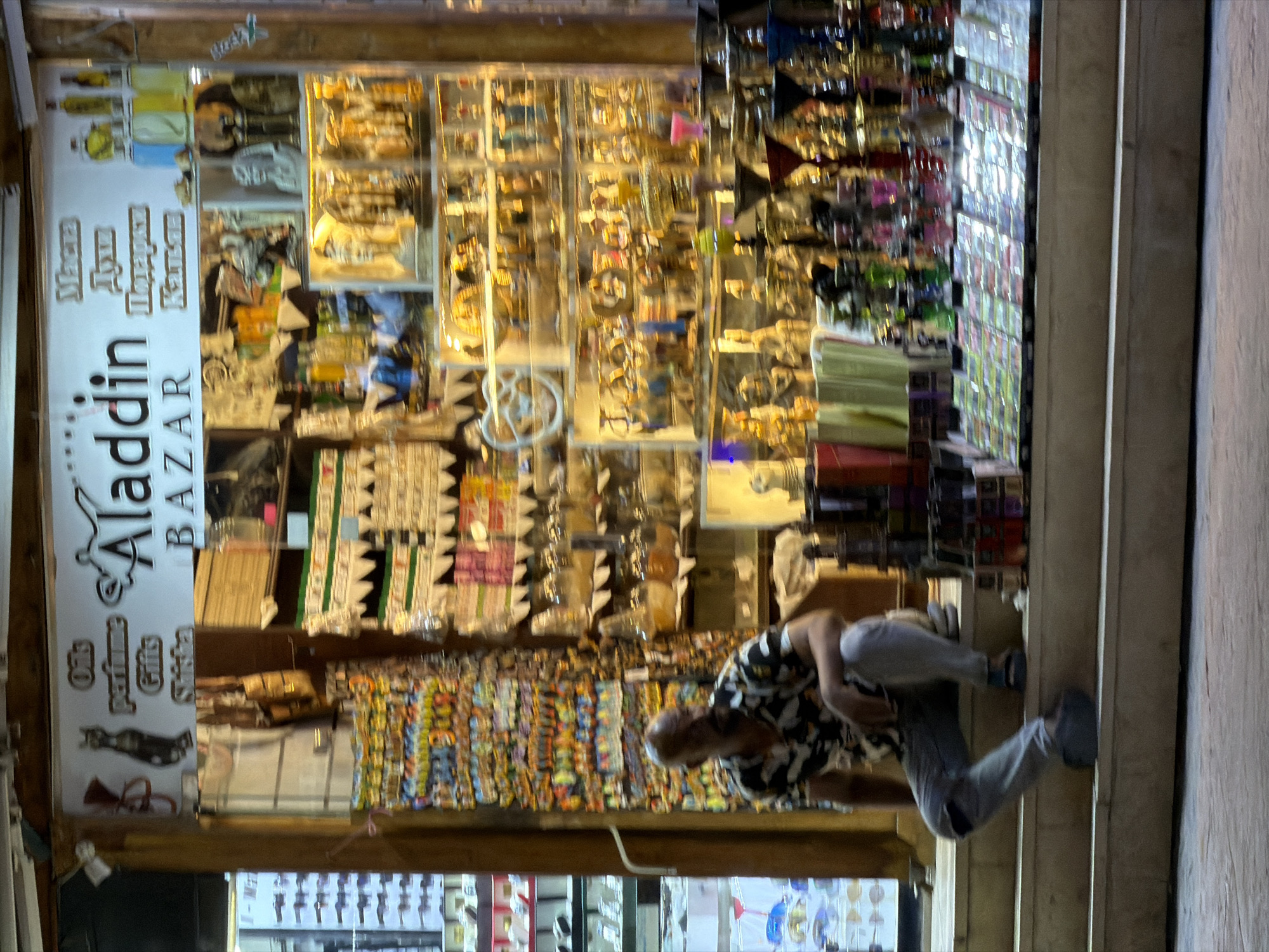 The image depicts the entrance of a souvenir shop named "Aladdin BAZAR" located in Sharm El Sheikh, Egypt. The shop's facade is filled with various items for sale, including oils, perfumes, gifts, and shisha. A man is seated on the steps leading up to the shop, wearing a patterned shirt and grey pants. The shop is brightly lit from within, showcasing a wide array of merchandise such as decorative statues, bottles of colored liquids, and numerous hookahs. The overall scene suggests a bustling market environment, typical of tourist areas in Egypt.