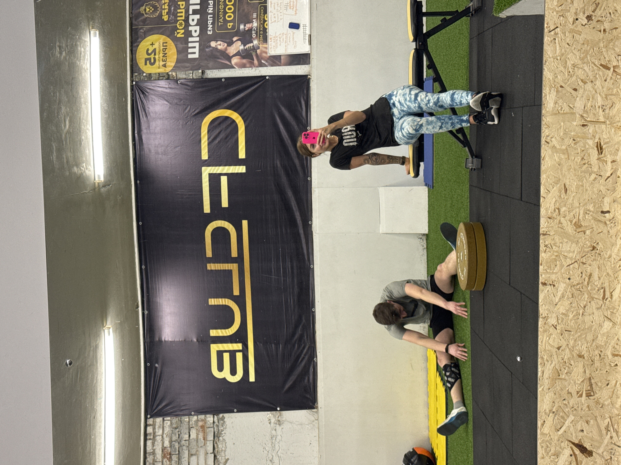 The image is an interior shot of a gym, likely in the city of Chaikovsky, Russia. On the left, there's a large black banner with the words "CLCrNB" in gold lettering. Above the banner is another sign or poster with Russian text.
A woman is visible in the upper right portion of the frame, standing on a workout machine and taking a photo of herself with a pink phone. She's wearing a black t-shirt and patterned leggings.
In the lower right portion of the image, a man is stretching on the black rubber gym flooring. He's wearing a gray t-shirt and black shorts, with a brace on his lower left leg. A weight plate sits on the floor next to him. The background wall is painted in neutral tones, and to the right is a wall of plywood. The floor is covered with black rubber mats.