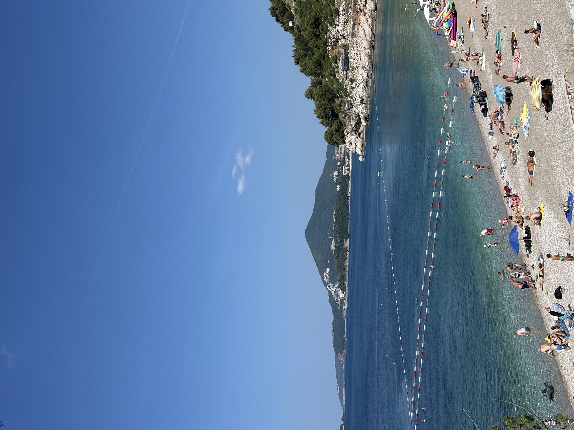 The image depicts a crowded pebble beach in Boreti, Montenegro, under a clear blue sky. The foreground is filled with sunbathers, some lying on towels, others sitting or standing. Beach umbrellas and loungers are scattered across the scene, along with some colorful inflatable toys. The shallow, clear turquoise water laps gently onto the shore, where several people are wading or swimming. Red and white buoys mark off a swimming area in the sea. In the background, a rugged, tree-covered coastline extends along the bay, with buildings dotting the hillsides. A large, cone-shaped mountain dominates the distant horizon. The scene conveys a typical summer day at a popular tourist destination.