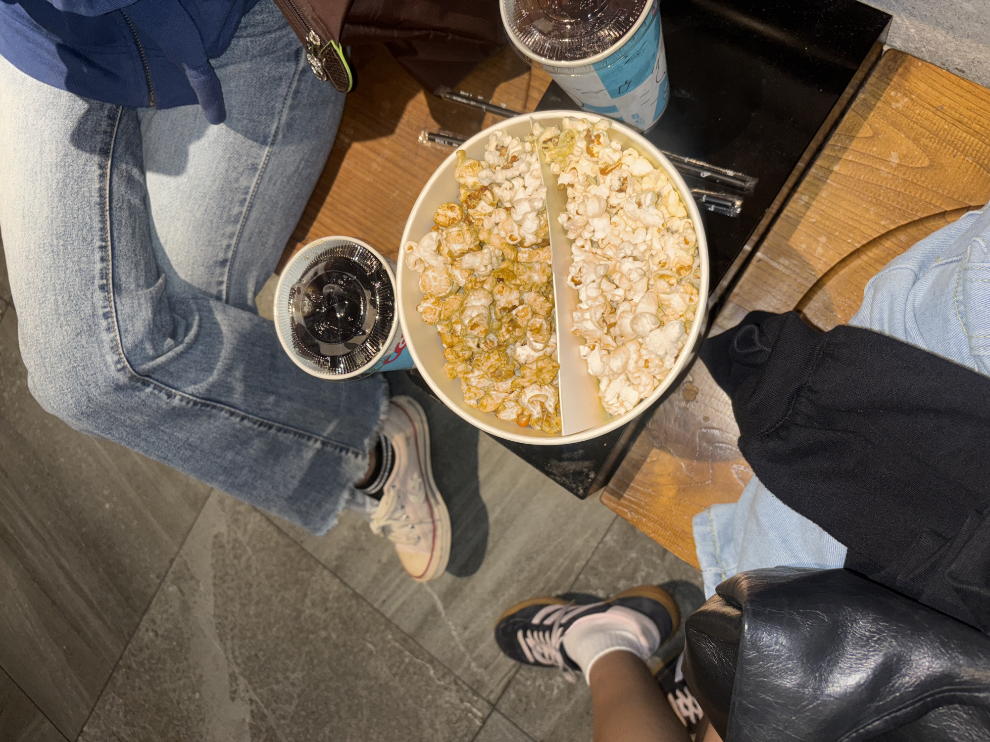 In West Jakarta, Indonesia, a scene unfolds with two individuals partially visible, seated on wooden furniture, sharing a large, divided bucket of popcorn and two drinks. One person, wearing blue jeans with frayed hems, has light-colored sneakers with pink accents. Another person, seen from the knee down, wears black athletic shoes with white socks. The environment suggests an indoor setting, possibly a cinema or entertainment venue, with a dark surface and tiled floor. One drink is a clear cup with a dark beverage and a lid, while the other is a blue and white cup with a similar lid. A black leather bag and a black hoodie are also present on the seating area, adding to the casual atmosphere. The popcorn is divided into two sections, one appearing to be caramel-colored, and the other plain. The lighting is dim, creating a cozy ambiance.