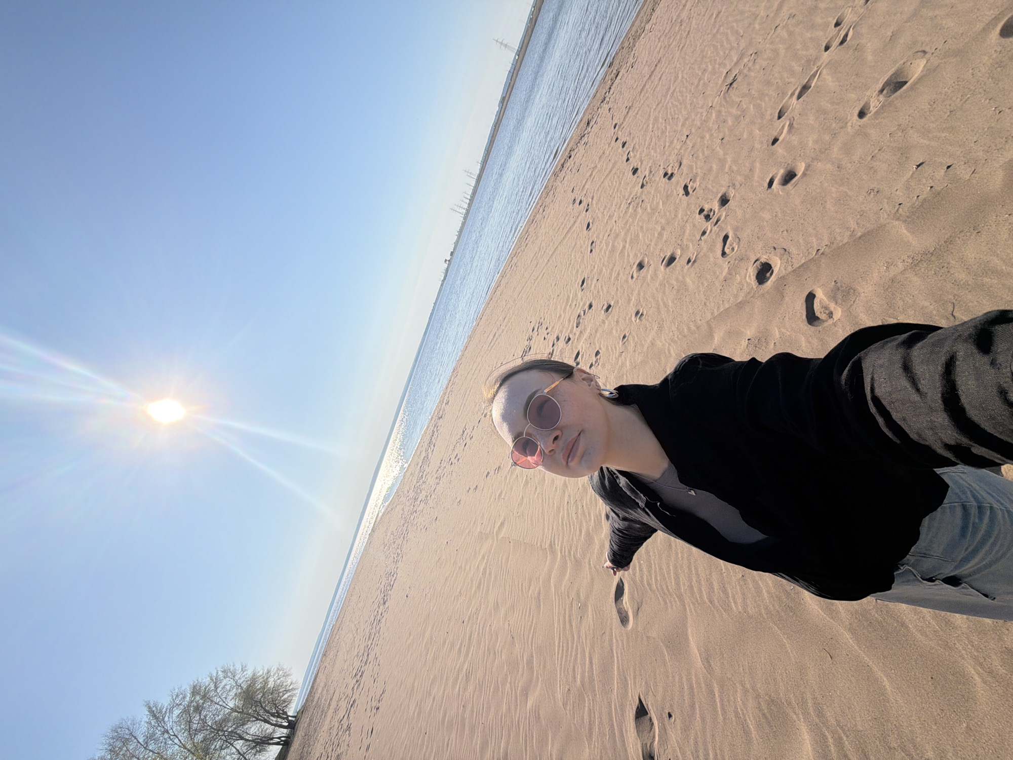 A person is standing on a sandy beach next to a body of water under a clear, sunny sky. The person is wearing sunglasses, a black jacket, and blue jeans. The beach has numerous footprints in the sand. In the distance across the water, there are industrial structures and power lines. Trees are visible in the bottom left corner. The sun is bright and high in the sky, causing glare. The location is Noviy, Russia.