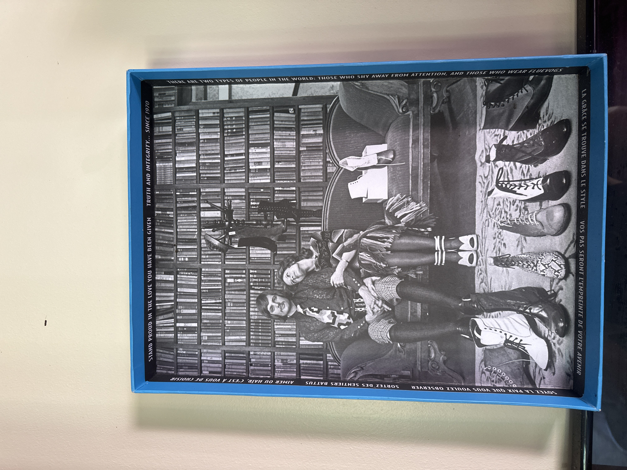 The image shows a framed poster with a black and white photograph of a man and a woman sitting on a couch in front of a wall of bookshelves. The man is wearing a patterned shirt and tie, and plaid pants. The woman is wearing a patterned dress. They are both looking at the camera. There are several pairs of shoes arranged on the floor in front of them. The poster has text in English and French around the edges. The poster is framed in a blue border. The image is cropped to show the poster, and the background is a plain wall. The location is not specified in the image.