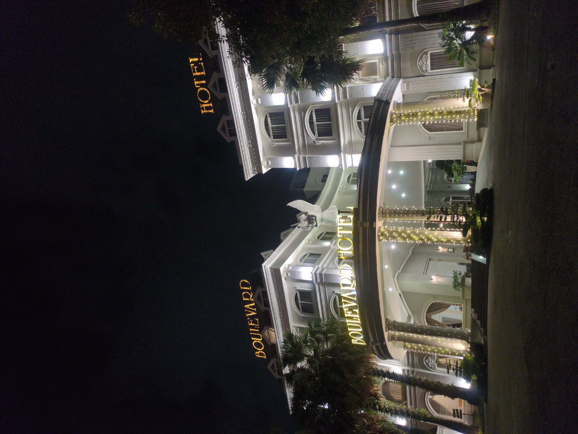 At night, the Boulevard Hotel in Phú Quốc, Vietnam, is illuminated. The building is white with an arched entrance and columns adorned with string lights. Palm trees are visible in front of the hotel. The words "BOULEVARD HOTEL" are displayed in illuminated letters on the front of the building. "HOTEL" is written in vertical gold letters. The sky is dark, indicating nighttime. The pavement is visible in the foreground, suggesting the location is an access road.