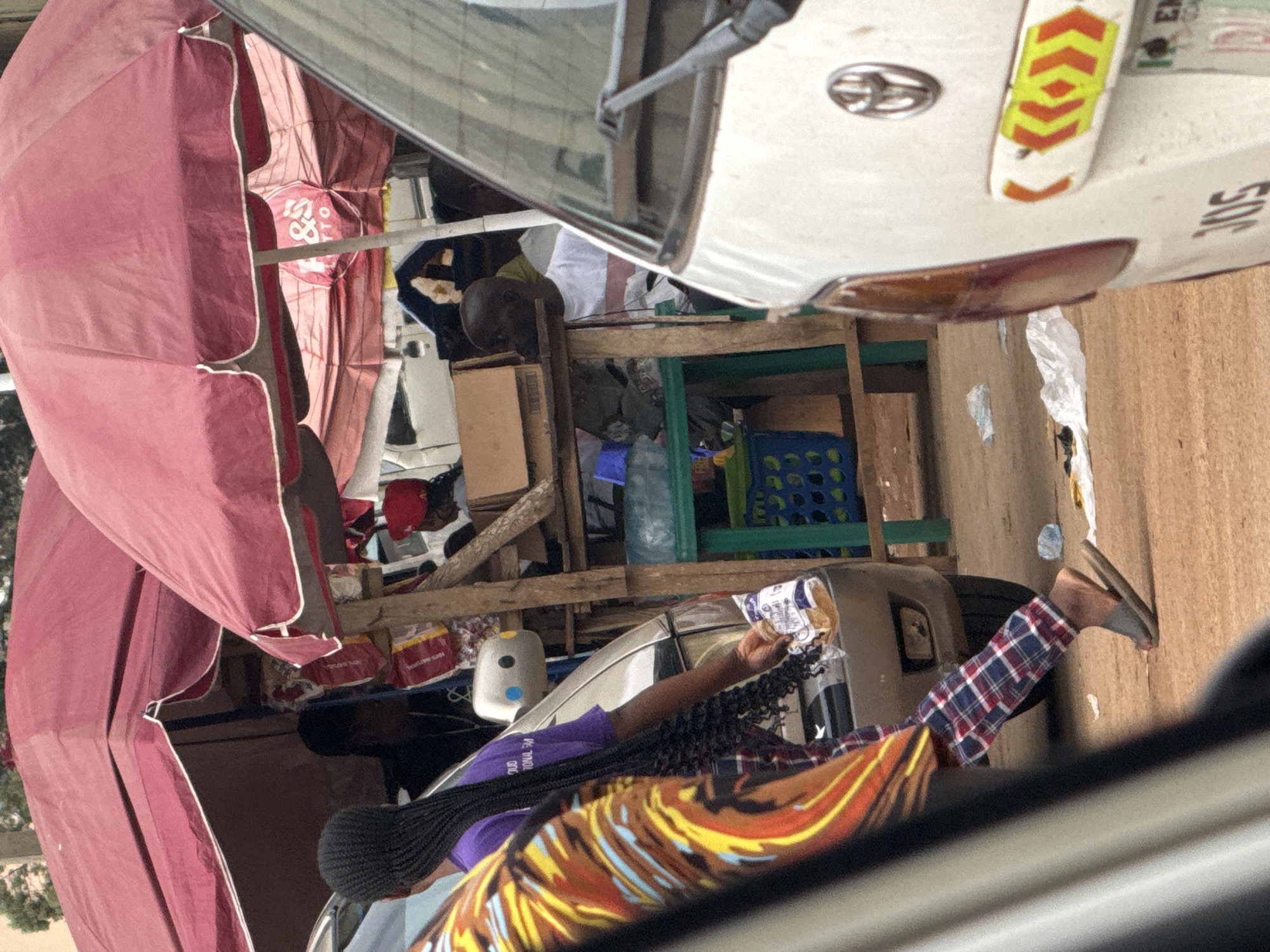 The image depicts a street scene in Orji, Nigeria, likely a marketplace or roadside stall area. Two large, red umbrellas provide shade over a wooden structure. Underneath the umbrellas, a stall is set up with various items, including packaged goods and boxes. Several individuals are visible, some obscured by the umbrellas and vehicles. One person in the foreground, wearing a purple shirt and plaid pants, walks with a plastic bag in their hand. Another person, seen from behind, has long braided hair. A white van is parked in the foreground, with its rear visible, displaying reflective safety markings. The ground is covered in reddish-brown dirt, with some scattered plastic debris. The overall impression is a busy, informal commercial setting.