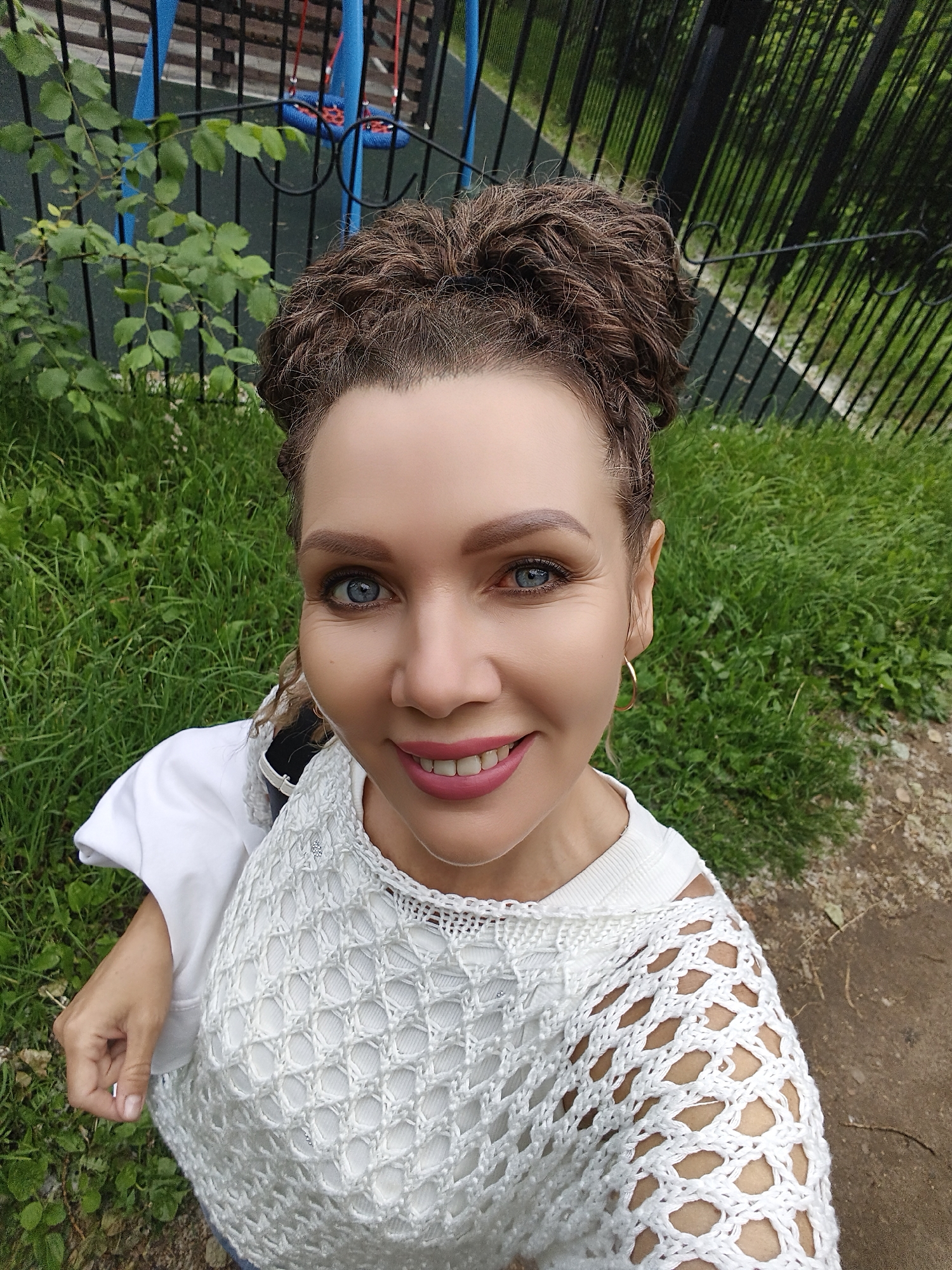 A medium shot photograph shows a woman outdoors, with her face filling much of the frame. She has brown, curly hair pulled up into two buns and blue eyes. She is smiling, showing her teeth, and wearing a white top with a crocheted overlay. In the background, there is green grass, a black metal fence, and what appears to be playground equipment. The overall scene suggests an urban park setting.