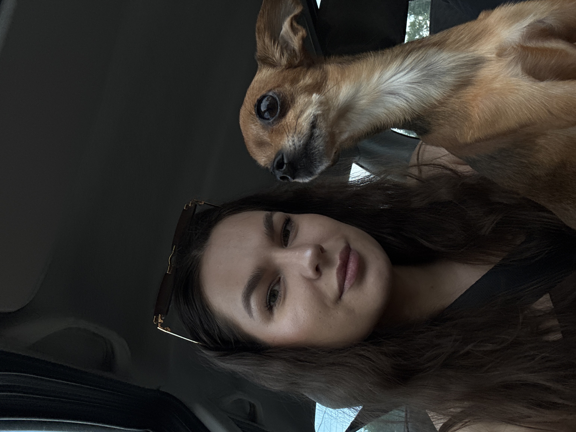 A young woman with long, dark hair and sunglasses perched on her head is in the driver's seat of a car. To her right, a small, tan dog with large, dark eyes is looking upwards and to the right. They appear to be traveling on a road near Rusalevka, Russia, with greenery visible outside the car windows. The woman is looking towards the camera with a slight smile. The scene is captured from a selfie perspective.