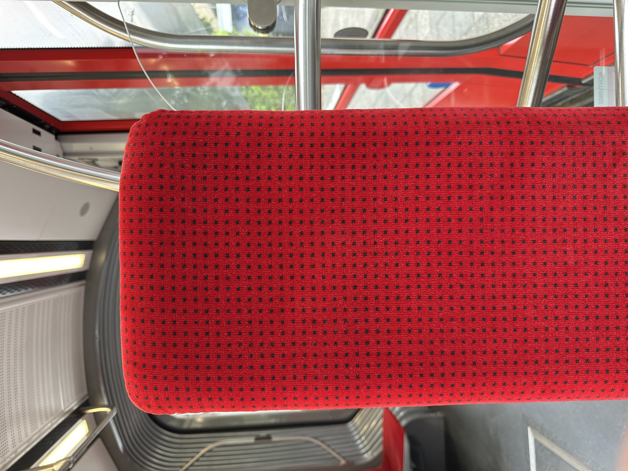 The image shows the red, patterned upholstery of a seat inside a cable car. The seat is the primary subject, filling most of the frame. The fabric has a repeating pattern of small, dark squares on a bright red background. To the right of the seat, a metallic railing is visible, along with a large window that offers a blurred view of the outside. Through the window, hints of green foliage and the red exterior of the cable car structure can be seen. To the left of the seat, the interior of the cable car is visible, including a curved, metallic wall and a glimpse of another red seat further back. The ceiling features fluorescent lighting. The image is a close-up interior shot, focused on the textures and colors of the cable car's seating. This cable car is part of the Seidenweberhaus cable car system in Krefeld, Germany.