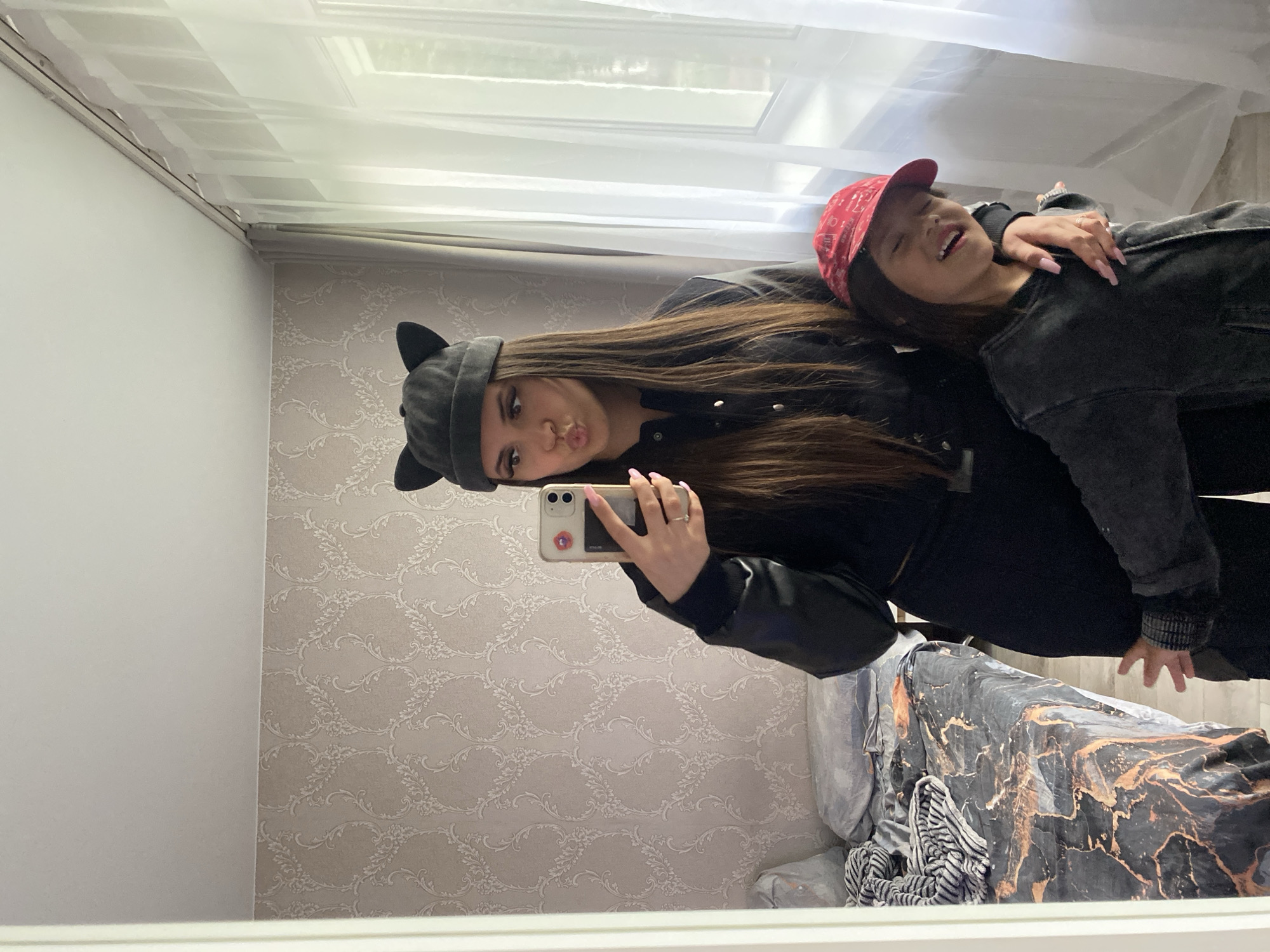 The image displays a full-body mirror selfie of two individuals in what appears to be a residential room. The foreground shows a young woman and a child, reflected in a mirror with a white frame on the left.
The young woman, positioned centrally, has long, straight, dark brown hair and is wearing a dark grey or black beanie with prominent cat ears. Her attire consists of a dark-colored jacket, possibly black, with long sleeves and light-colored snaps or buttons down the front. She is holding a light-colored smartphone with a dark case in her left hand, which has light pink polished fingernails, towards the mirror to capture the image. A ring is visible on her left ring finger. She is making a "puckered lips" expression towards the camera.
To her right, partially embraced by the woman, is a child wearing a red baseball cap and a dark, possibly charcoal grey, textured jacket. The child is looking upwards and to the right, with eyes partially closed, exhibiting a joyful expression, possibly a laugh or a smile. The young woman's right hand rests on the child's left shoulder.
The room's environment includes a wall adorned with light-colored, possibly beige or taupe, patterned wallpaper featuring a repetitive, ornate, floral or damask design. Above the wallpaper, the ceiling is plain white. To the left, a bed with a dark grey fitted sheet featuring abstract orange or copper-colored patterns is visible. A light grey pillow and a striped blanket are present on the bed. To the right, a large window provides natural light, covered by sheer white curtains suspended from a white curtain rod. An opaque, light-colored curtain is visible to the left of the sheer curtains. The specific city of Chaikovsky, Russia, cannot be determined from the visual information within the image.
