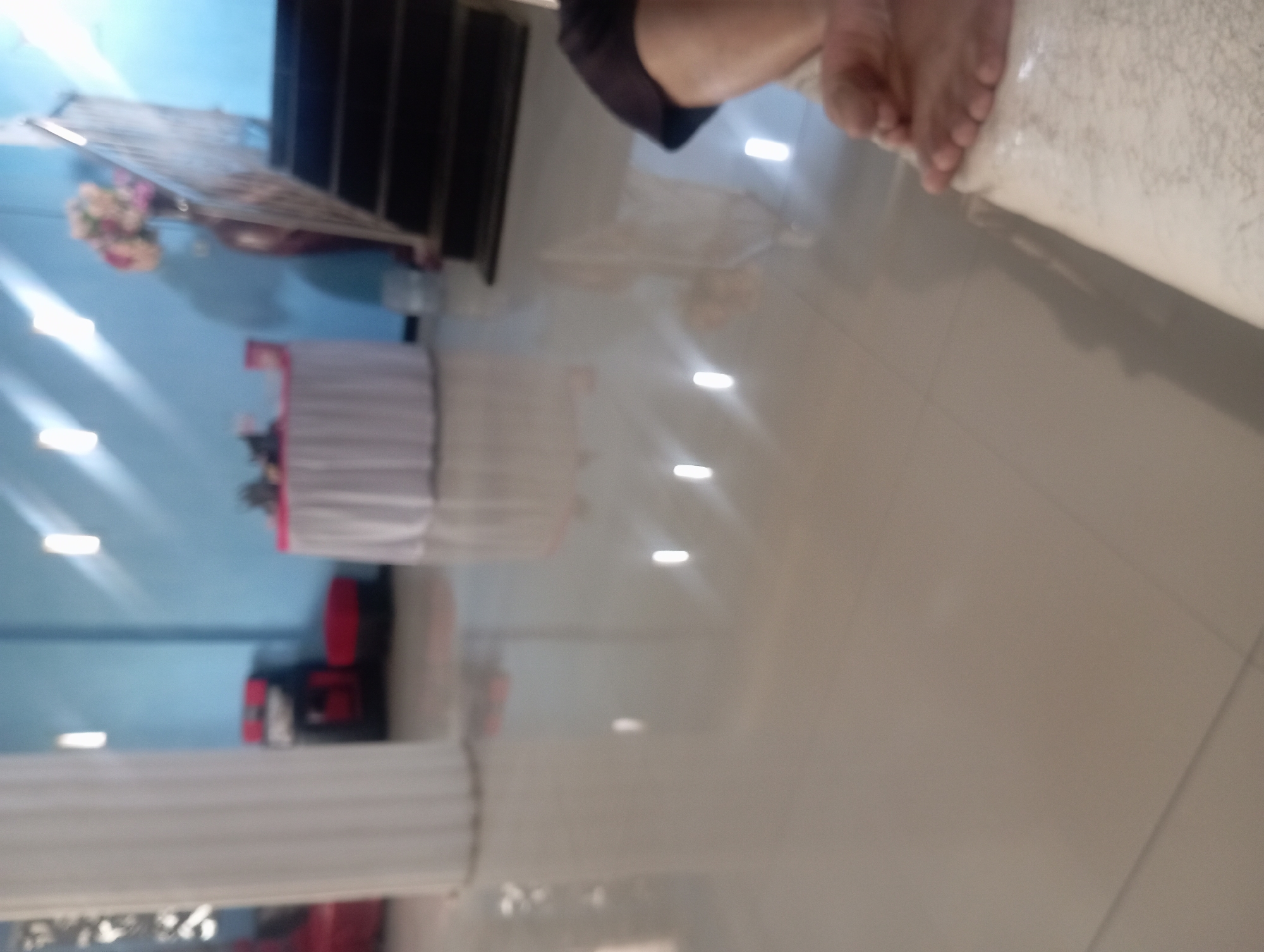 A person's feet are visible in the foreground of this blurry indoor image, resting on a light-colored surface. The background reveals a room with light blue walls, a white fluted pillar, a table draped with a white tablecloth, and a set of dark stairs. There are also decorative elements like a vase with flowers and some red furniture. The overall impression is of an interior space, possibly a hall or a room for events, located in Kudus, Indonesia. The lighting suggests it is daytime or well-lit indoors.