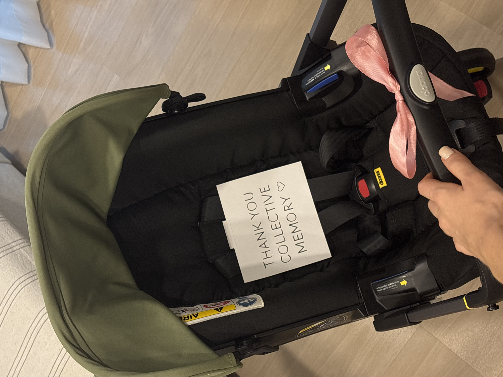 A top-down, indoor shot focuses on a Doona baby car seat resting on a tan-colored floor, possibly in a home. The car seat is primarily black, with an olive green canopy extending from the left side. A white card is placed in the seat, with the text "THANK YOU COLLECTIVE MEMORY♡" printed on it in gray letters. A pale pink ribbon is tied around the black handle of the car seat. A fair-skinned hand with manicured nails is seen holding the handle on the right side of the frame. The floor appears clean and smooth. A partial view of a striped sofa and some white material are on the left. The image is well-lit.