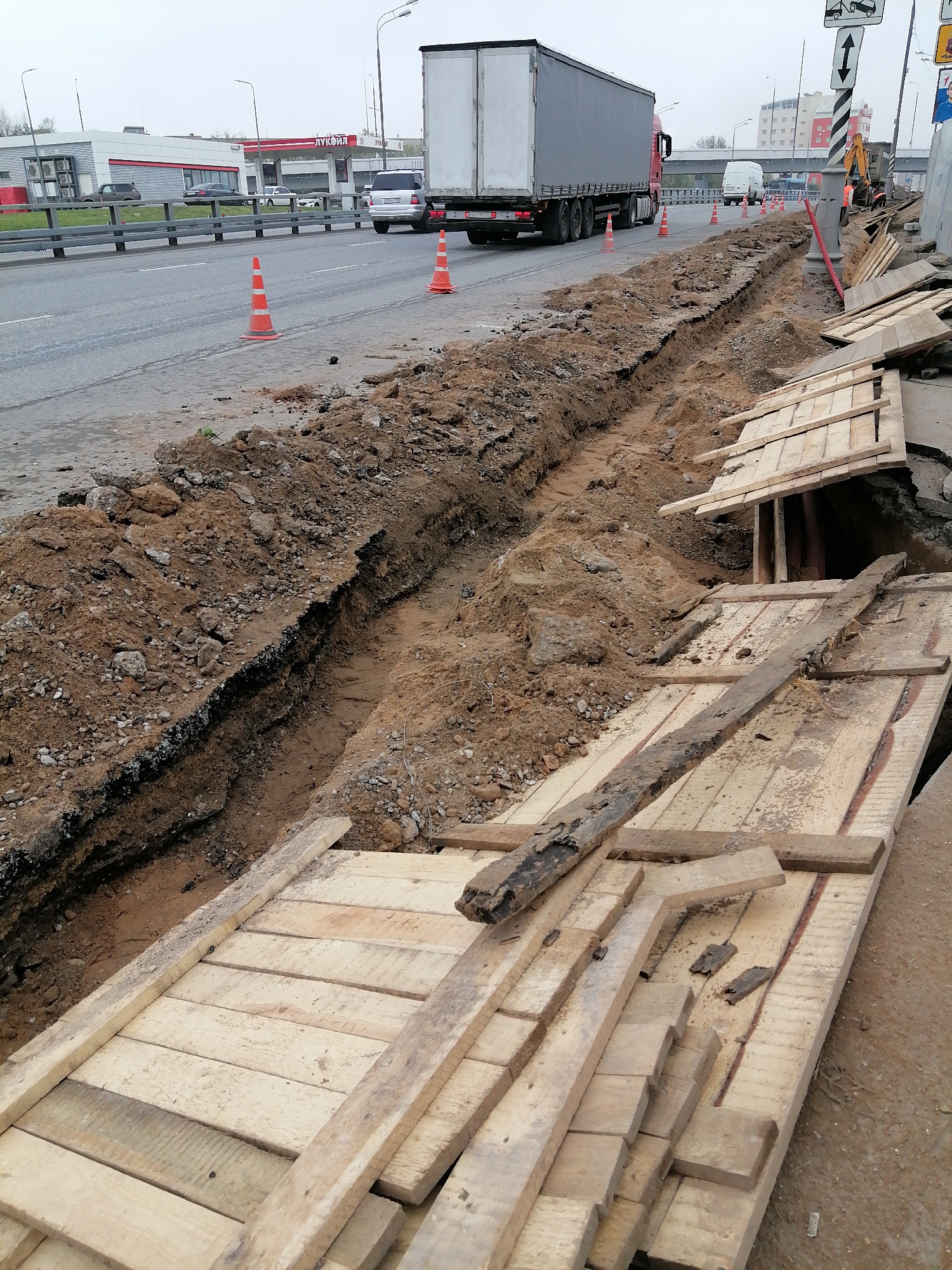 The image depicts an infrastructure project in Moscow, Russia, where a trench has been excavated along a road. The trench is bordered by dirt and asphalt, with wooden planks laid across it for support and possibly pedestrian or equipment access. A large truck drives along the road, indicated by orange cones marking the construction zone. In the background, there is a "Лукойл" (Lukoil) gas station and a building with multiple air conditioning units. An excavator is visible further down the road. The day appears overcast.