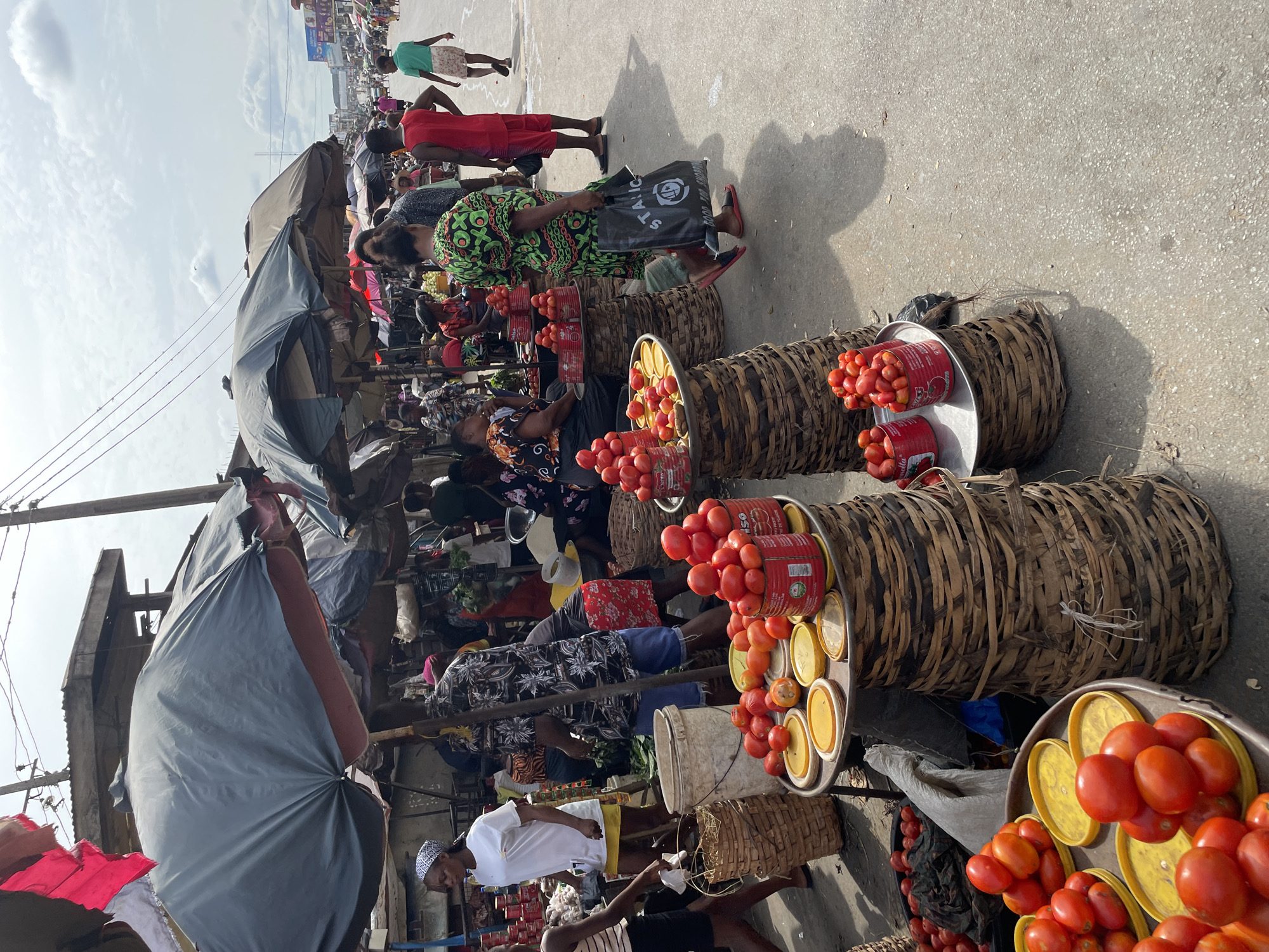 The image depicts a bustling outdoor market in Onitsha, Nigeria. Several vendors are seated behind large, round woven baskets filled with tomatoes and canned tomato paste. Some tomatoes are piled on aluminum trays with yellow lids. Pedestrians are walking along a concrete street in the background. Some vendors are shaded by umbrellas and makeshift tarps. People are wearing a variety of clothing.