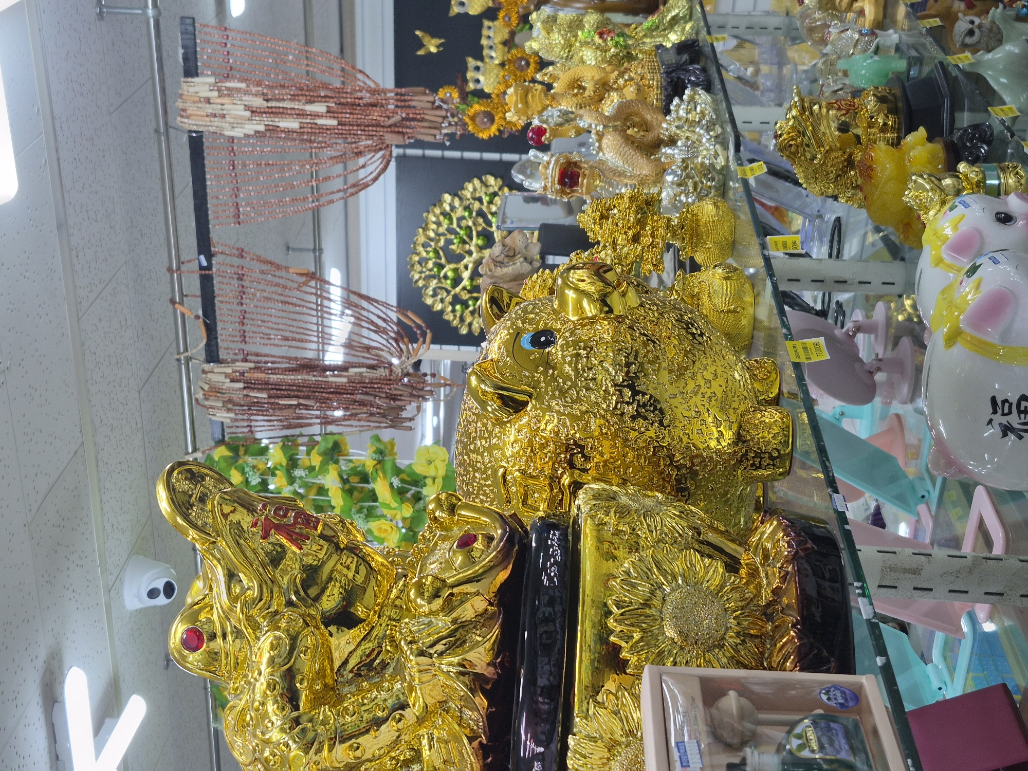 The image shows a shelf filled with various golden decorative items, likely in a store, in Ansan, South Korea. Dominating the foreground are two large, shiny golden piggy banks. To the left, a golden dragon-like figure is prominently displayed, with a red jewel for an eye and a red Chinese character, "福" (fú, meaning fortune), on its head. Behind the dragon, a beaded curtain hangs, and further back, a decorative golden tree with green accents is visible. Several other smaller golden figurines, including what appear to be owls and a dragon, are arranged on the shelf. A security camera is mounted on the ceiling. The overall impression is a collection of auspicious and decorative items commonly found in East Asian markets.