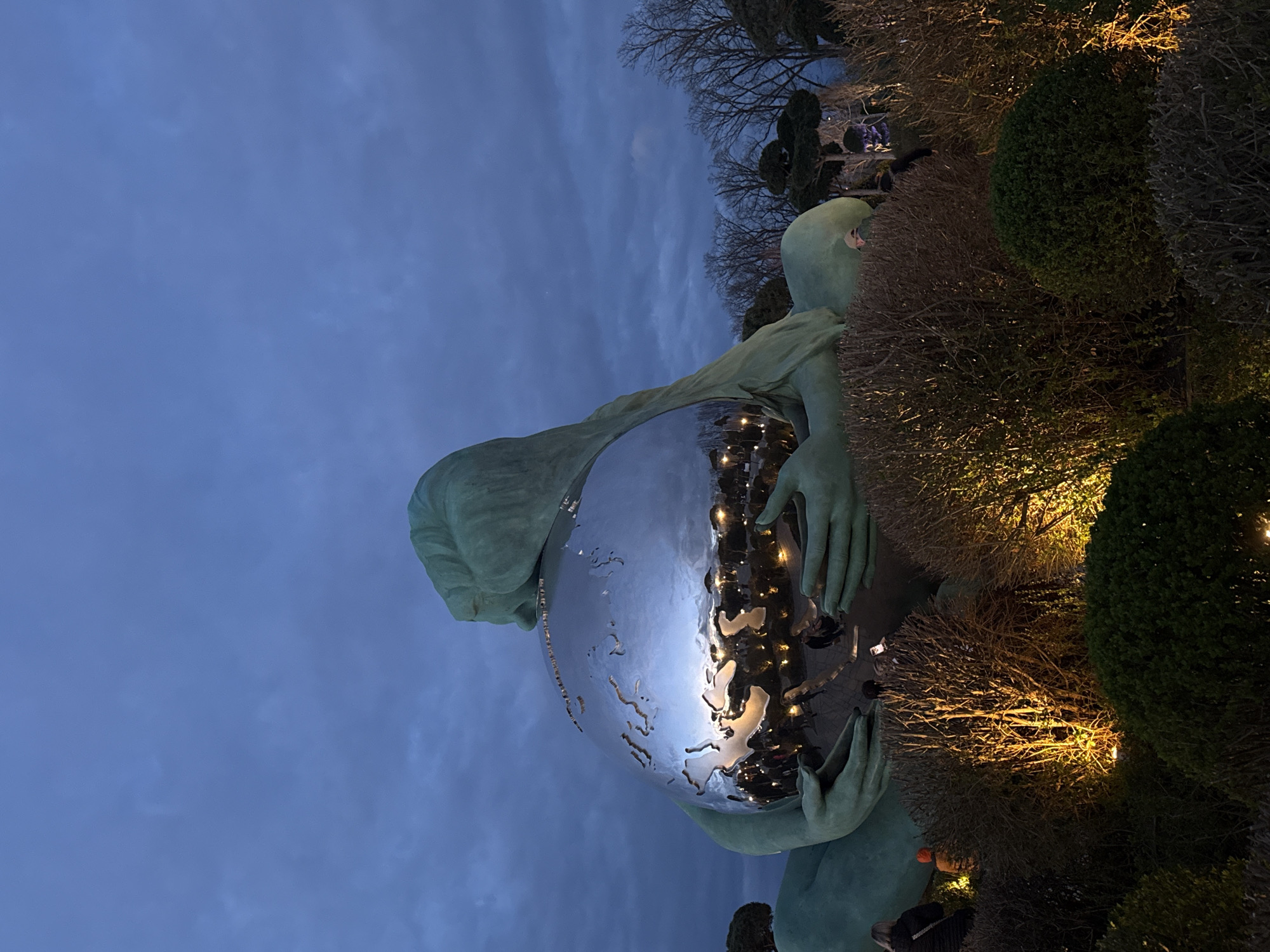 A low-angle, rotated photograph features a metallic sculpture of a person holding a reflective sphere. The statue is light green and appears to be made of stone or metal. The sphere reflects lights, shapes, and colors, suggesting the sculpture is located in a public space, possibly at night or dusk. The sky is visible in the background, colored in hues of blue and grey, indicating twilight. Shrubbery surrounds the base of the statue.