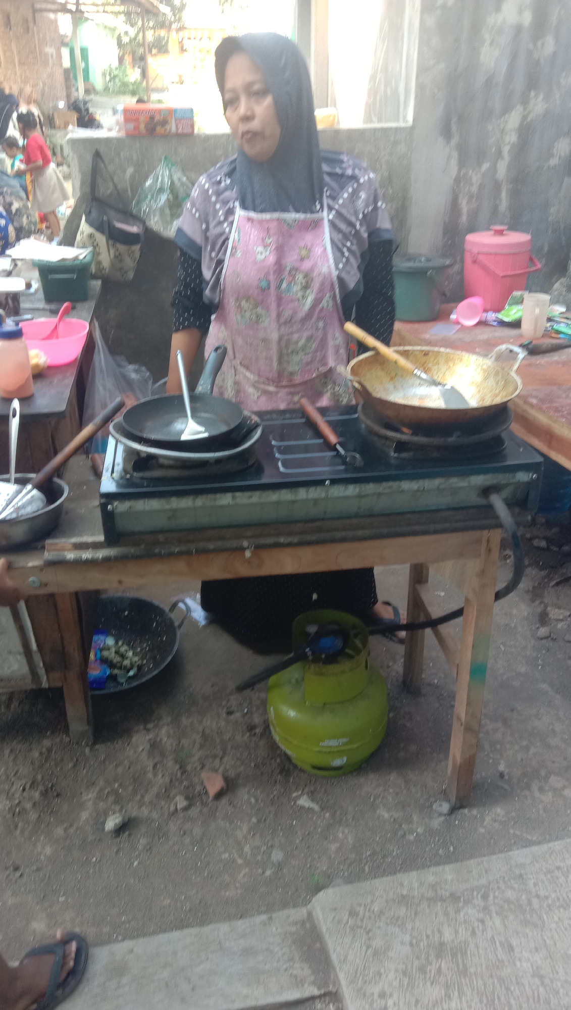 An Indonesian woman wearing a headscarf and an apron stands in front of a portable gas stove. The stove has two burners, with a frying pan on the left and a wok on the right. A green LPG gas cylinder is placed on the ground beneath the table supporting the stove. In the background, a pink cooler box and other items are visible. The scene appears to be an outdoor food stall, possibly in Karanganyar, Indonesia, given the context. The ground is dirt and gravel, and there are other indistinct figures and structures in the background, suggesting a bustling environment.