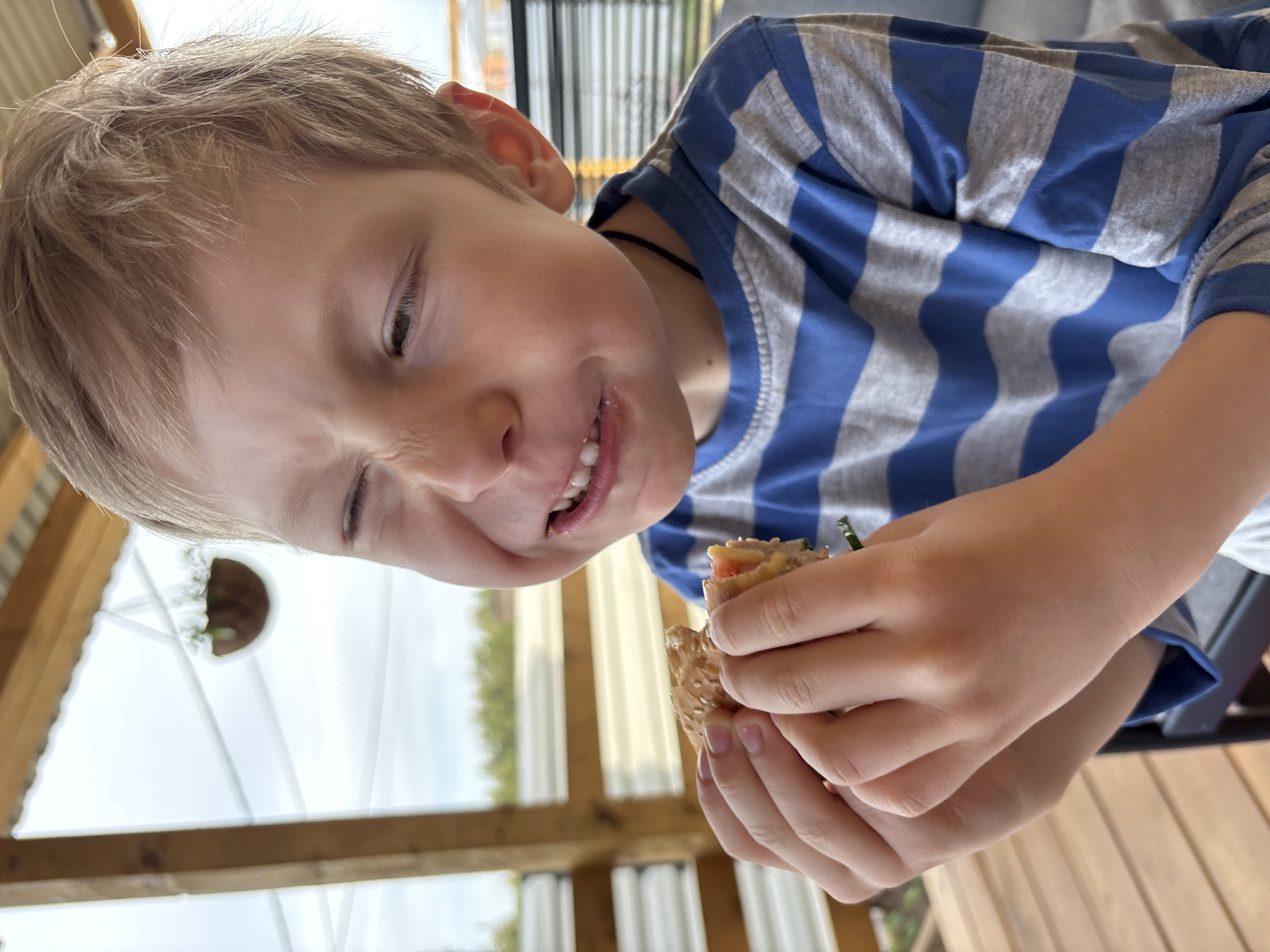 A child with blond hair wearing a blue and gray striped shirt is eating a sandwich. The child is holding the sandwich in both hands and appears to be making a face while eating. The background includes a wooden structure and a glimpse of outdoor scenery. The specific location within Chaikovsky, Russia, cannot be determined from the image.