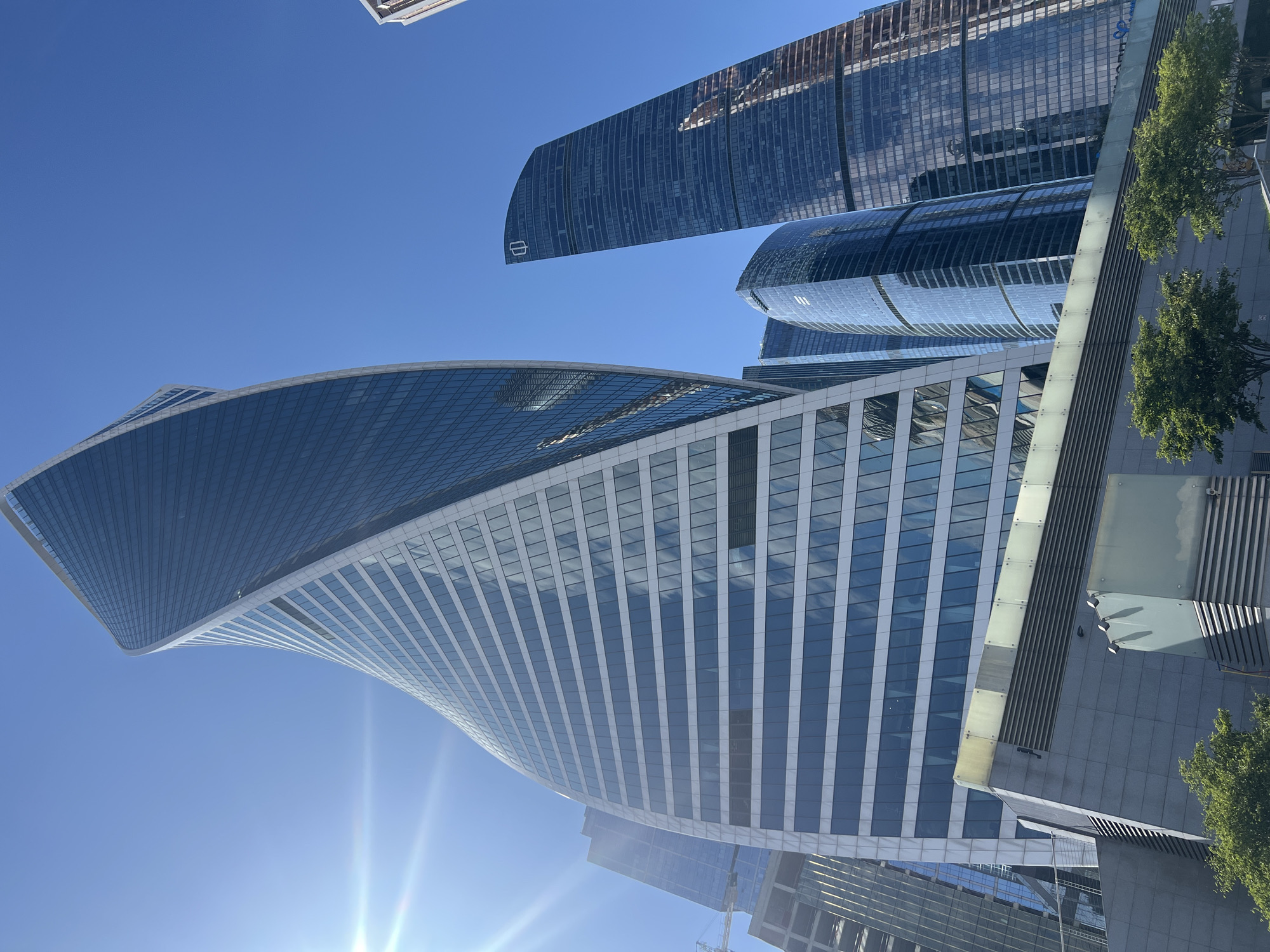 The image captures a low-angle view of several modern skyscrapers in Moscow City, Russia, under a clear blue sky. The most prominent building is the "Neva Towers 2," a skyscraper with a distinctive twisted facade, clad in glass and white panels. To its right, the "Eurasia Tower" is visible, a taller, slender skyscraper with a dark, reflective glass exterior. Other buildings in the Moscow City complex, such as the "Federation Tower" or "OKO Towers," can be seen in the background, also featuring modern architectural designs and glass facades. The sun is shining brightly, creating lens flare in the upper left corner and casting reflections on the buildings. In the foreground, a portion of a lower-level structure with grey panels and horizontal vents is visible, along with some green trees. There are no people or vehicles depicted in the image. The scene is static and presents a snapshot of the contemporary urban landscape of Moscow's financial district.