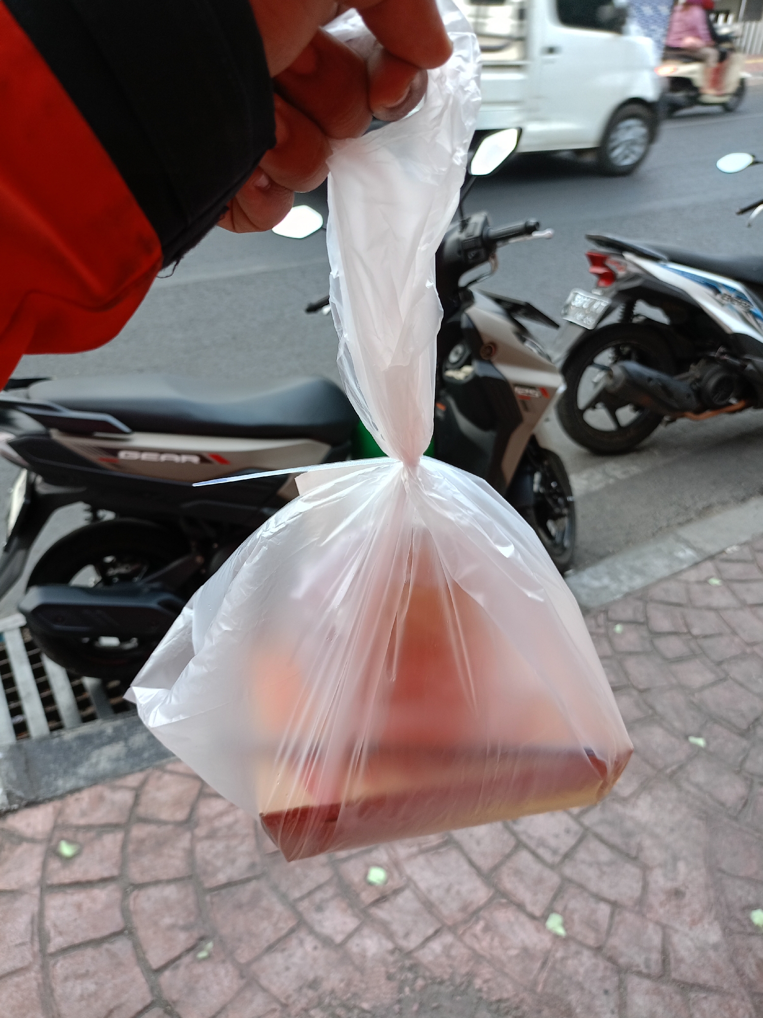 A person's hand, wearing a black and red jacket, holds a transparent plastic bag filled with a liquid and what appears to be food. The bag is tied at the top. In the background, a street scene in Batu, Indonesia is visible. There are several motorcycles parked and in motion, and a white truck is also present. The ground is paved with reddish-brown interlocking bricks. The overall lighting suggests it is daytime.