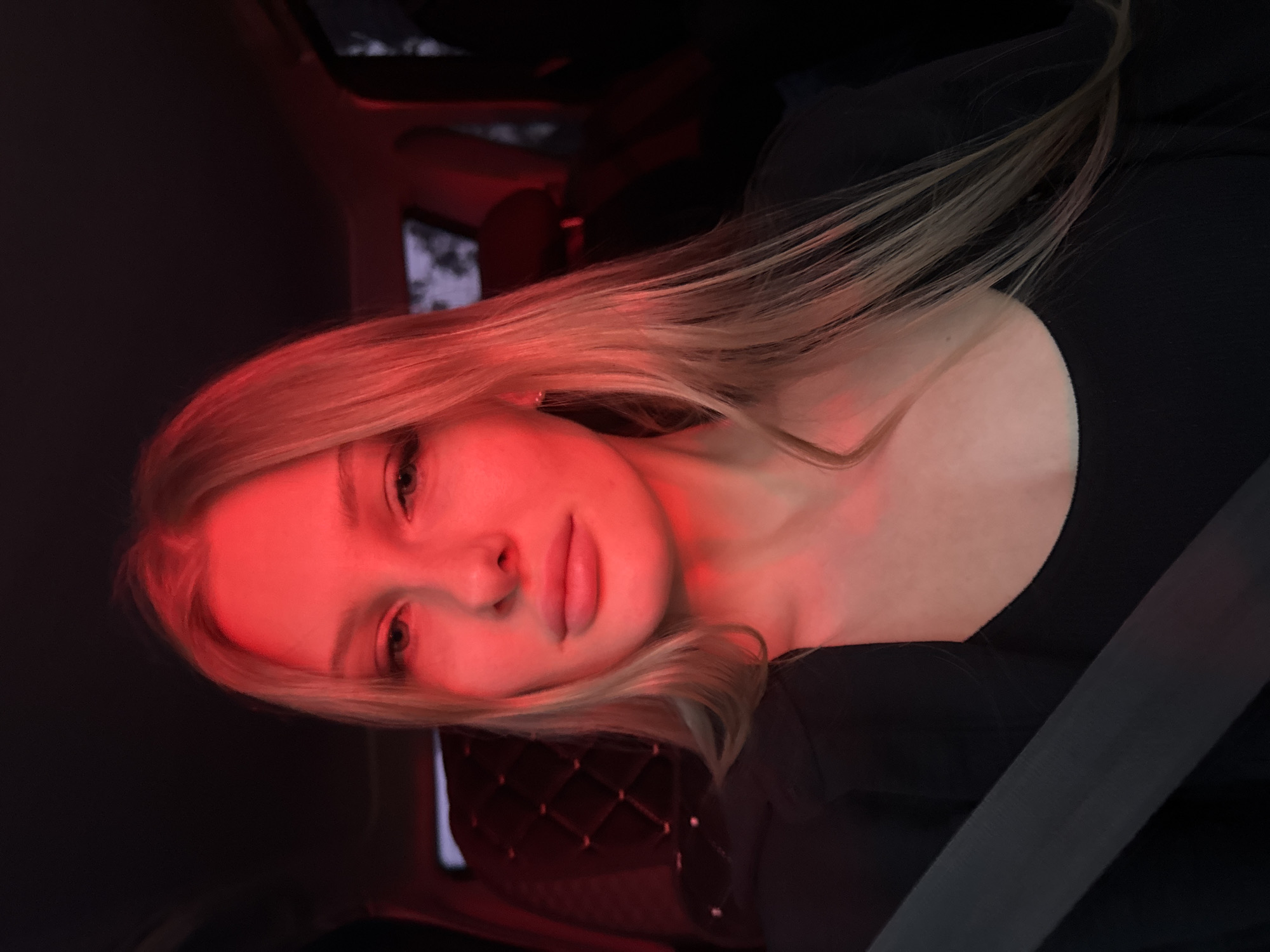 A young woman with blond hair sits in a car interior illuminated by red light. She is wearing a black top. A seatbelt is visible across her chest. The car appears to have diamond-patterned stitching on the interior panels. The specific location in the city of Perm, Russia cannot be determined from this image.