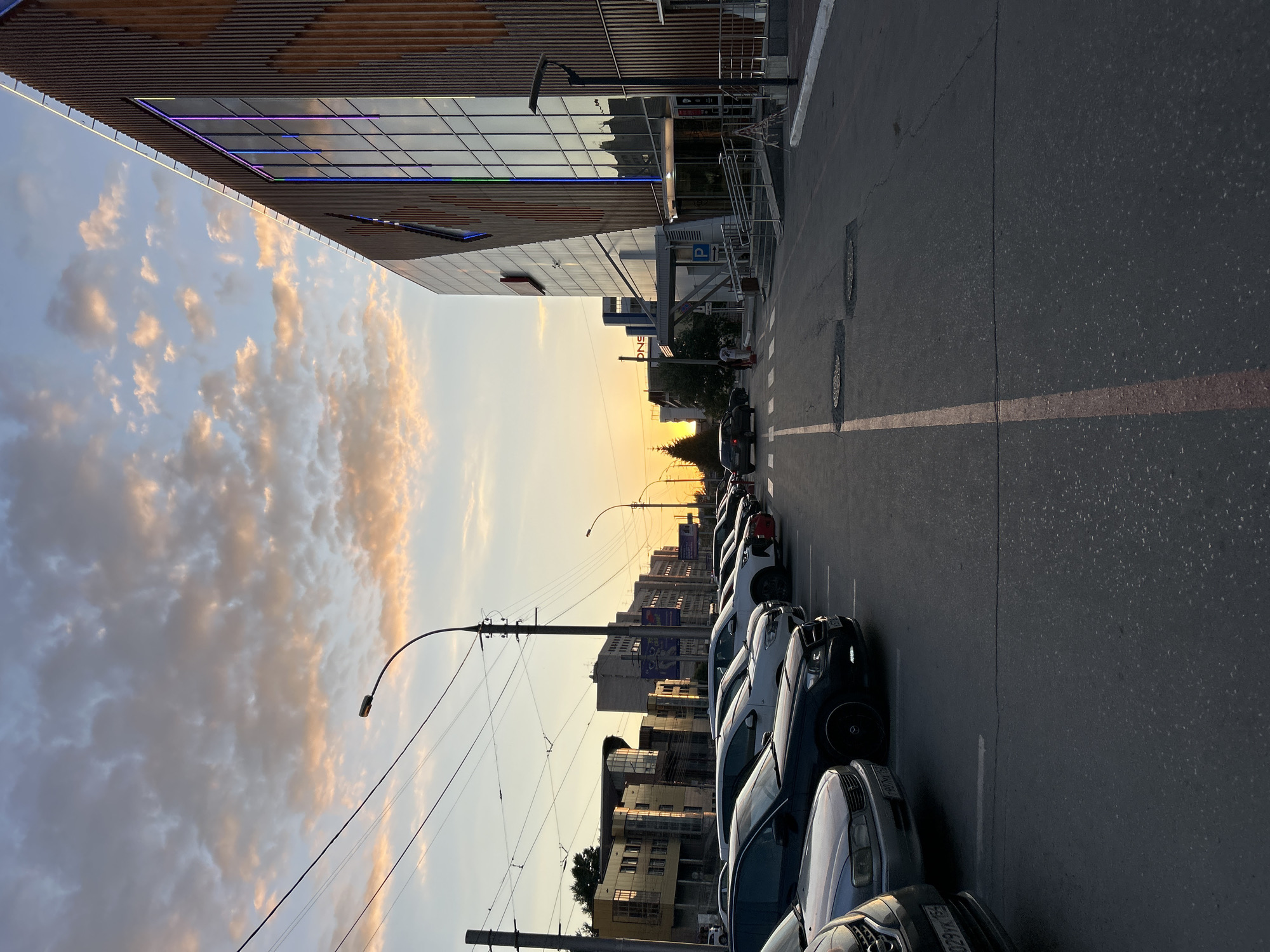 The image depicts a street scene in a city at dusk. The sky is filled with scattered clouds illuminated by the setting sun, casting a warm glow. On the right side of the frame, a modern building with a glass facade and wooden accents is visible, featuring colorful LED lighting. In front of the building, there is an entrance with a ramp and stairs. To the left, a row of parked cars lines the street. Beyond the cars, several multi-story buildings are seen in the background, along with streetlights and utility wires. The road surface appears to be asphalt with painted lane markings. A few people are walking on the sidewalk near the building entrance. The overall atmosphere suggests an urban environment during the evening transition.
