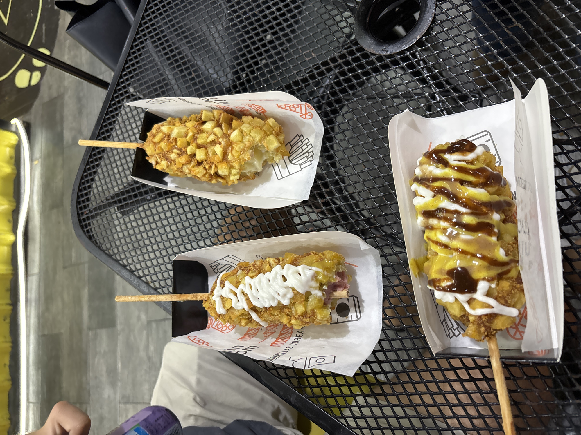 The image shows three Korean corn dogs on a black metal table. Each corn dog is on a stick and served in a white paper holder. One corn dog is coated in potato cubes and drizzled with a white sauce, another is coated in potato cubes only, and the last is coated in potato cubes and drizzled with yellow and brown sauces. The background includes a blurred glimpse of what appears to be a person’s hand and a beverage. Given the brand visible on the paper holder, the location is likely in Veracruz City, Mexico.
