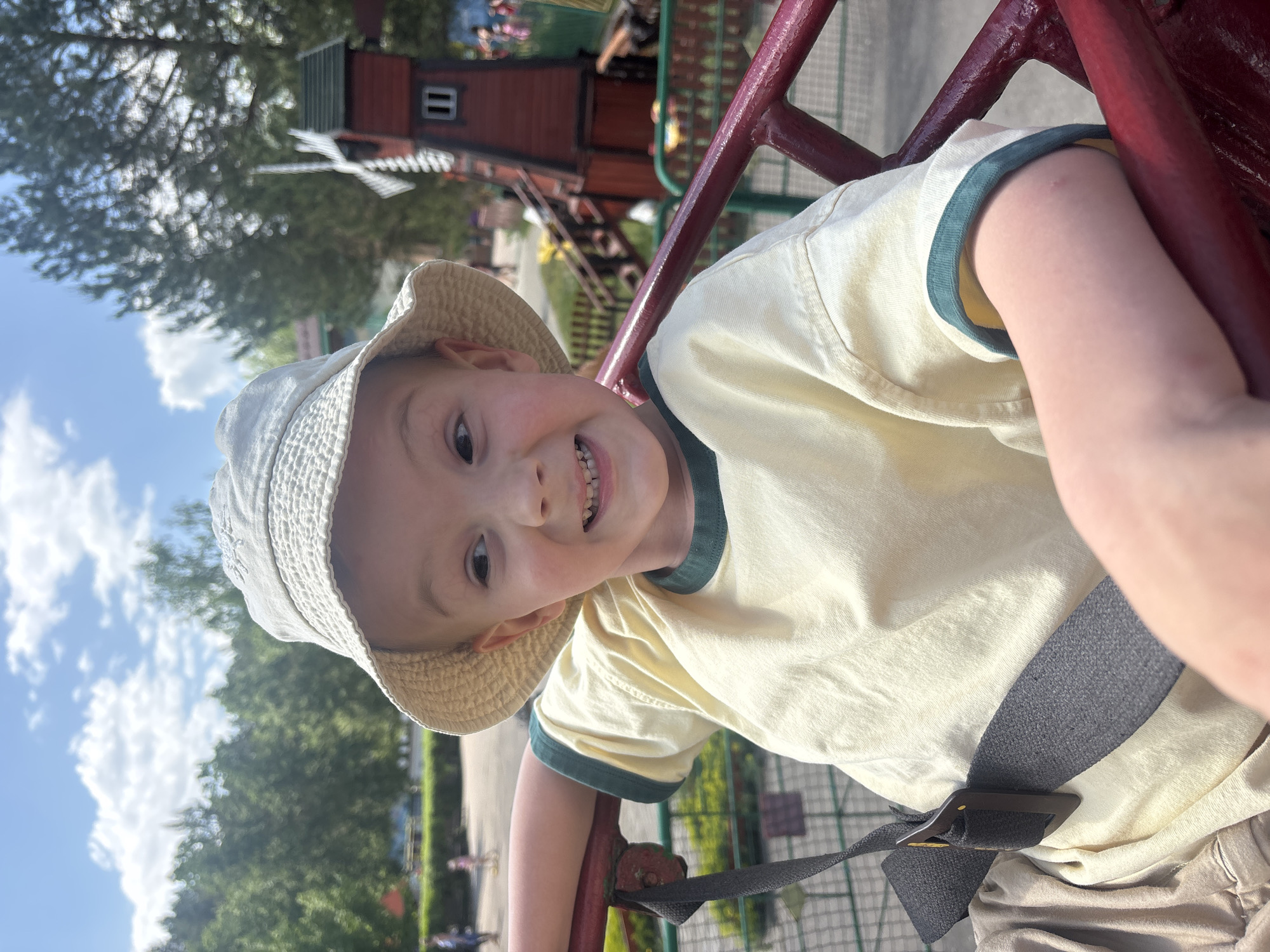 A young boy, wearing a light yellow t-shirt with dark green trim and a tan bucket hat, is secured in a ride with a grey safety strap and red metal frame. He is smiling and looking at the camera. In the background, there are trees, a partially visible red and brown building with a windmill, and other people. The location is the city of Chaikovsky, Russia.