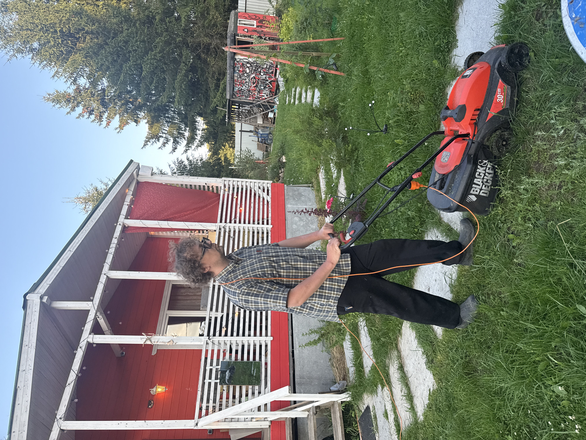 A man with curly hair and glasses, wearing a plaid shirt and black pants, is mowing the lawn in front of a red wooden house. He is pushing an orange and black electric lawnmower with a visible cord. The house has a white porch with a red railing and is decorated with string lights. The lawn is green and slightly overgrown in places. In the background, there are evergreen trees and a small shed covered in graffiti. The scene appears to be a residential yard in Lyady, Russia, on a clear day with soft lighting.