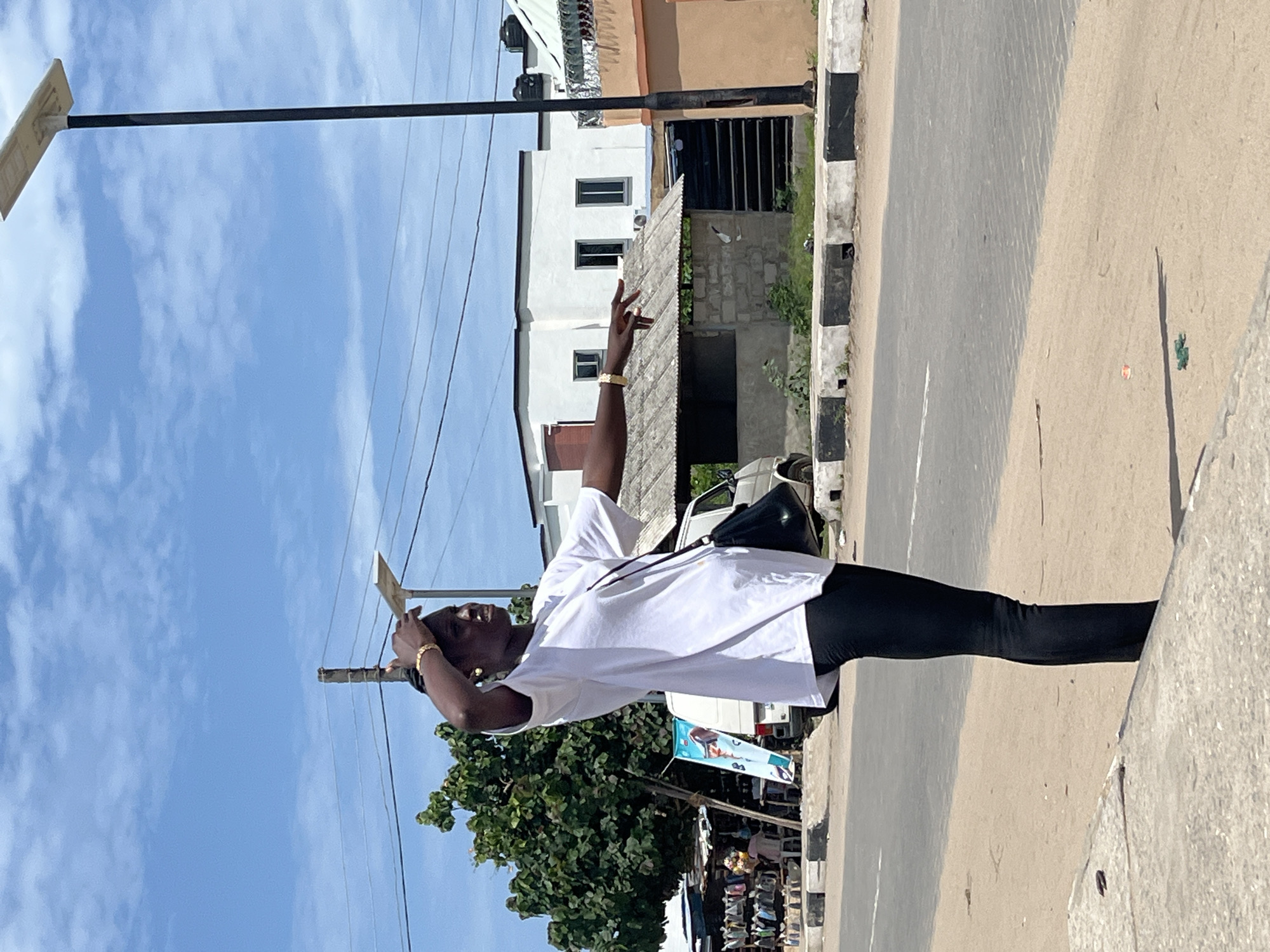 The image shows a person wearing a white t-shirt and black leggings, standing on a paved sidewalk in what appears to be Badagry, Nigeria, based on the general setting and architecture. The person is holding what appears to be a small, light-colored sign or object near their head, and their other arm is raised with fingers extended in a gesture. The background includes a street, buildings with white exteriors, a light pole, power lines, trees, and a row of street vendors or stalls. The weather appears to be sunny with a clear blue sky.