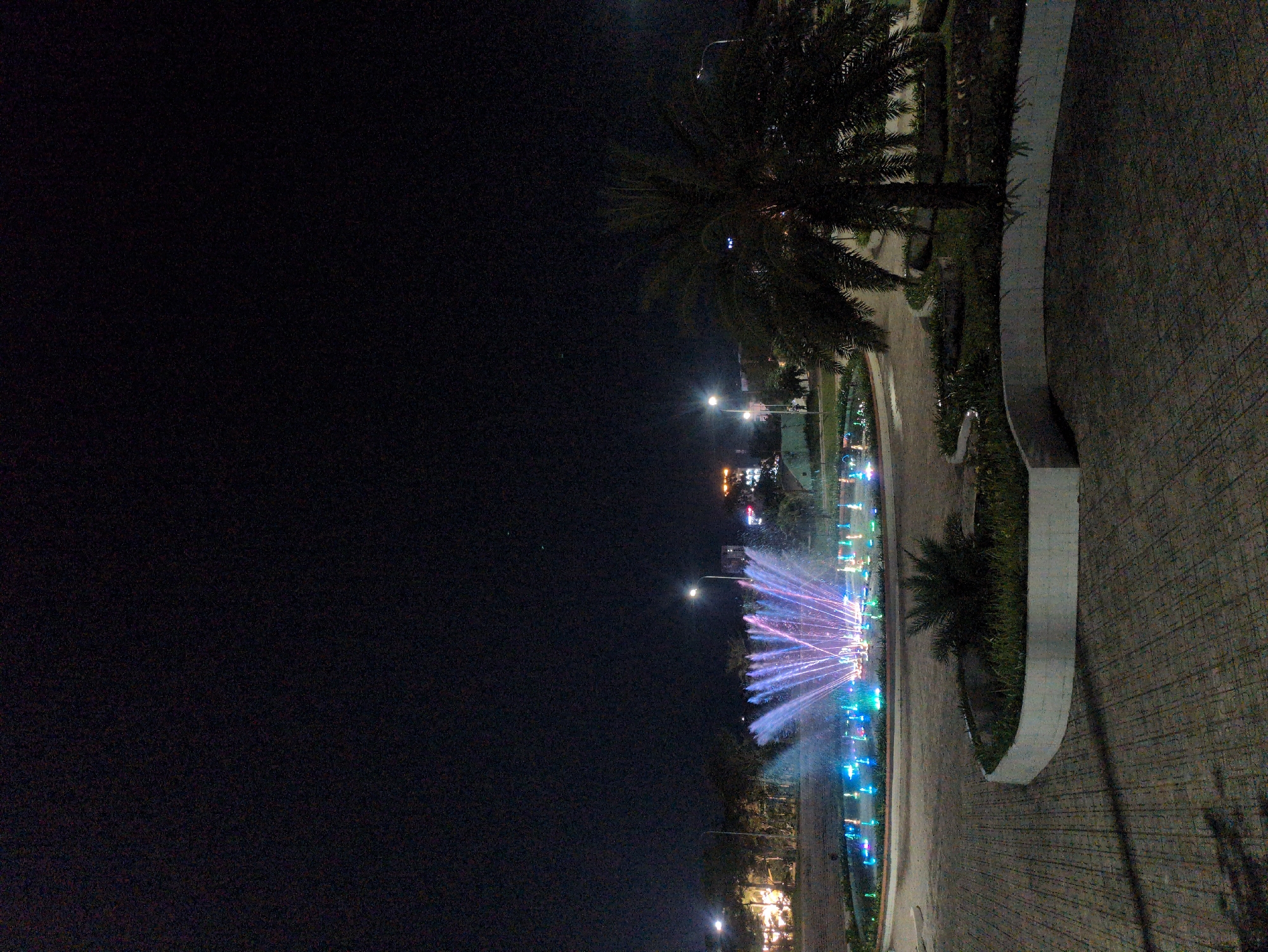 The image depicts a nighttime scene in Tuy Hòa, Vietnam. A brightly illuminated fountain is the focal point, displaying a vibrant array of blue, pink, and purple lights. The surrounding area includes palm trees and paved pathways. The sky is dark, and other buildings with lights can be seen in the distance. The photograph appears to have been taken from ground level.