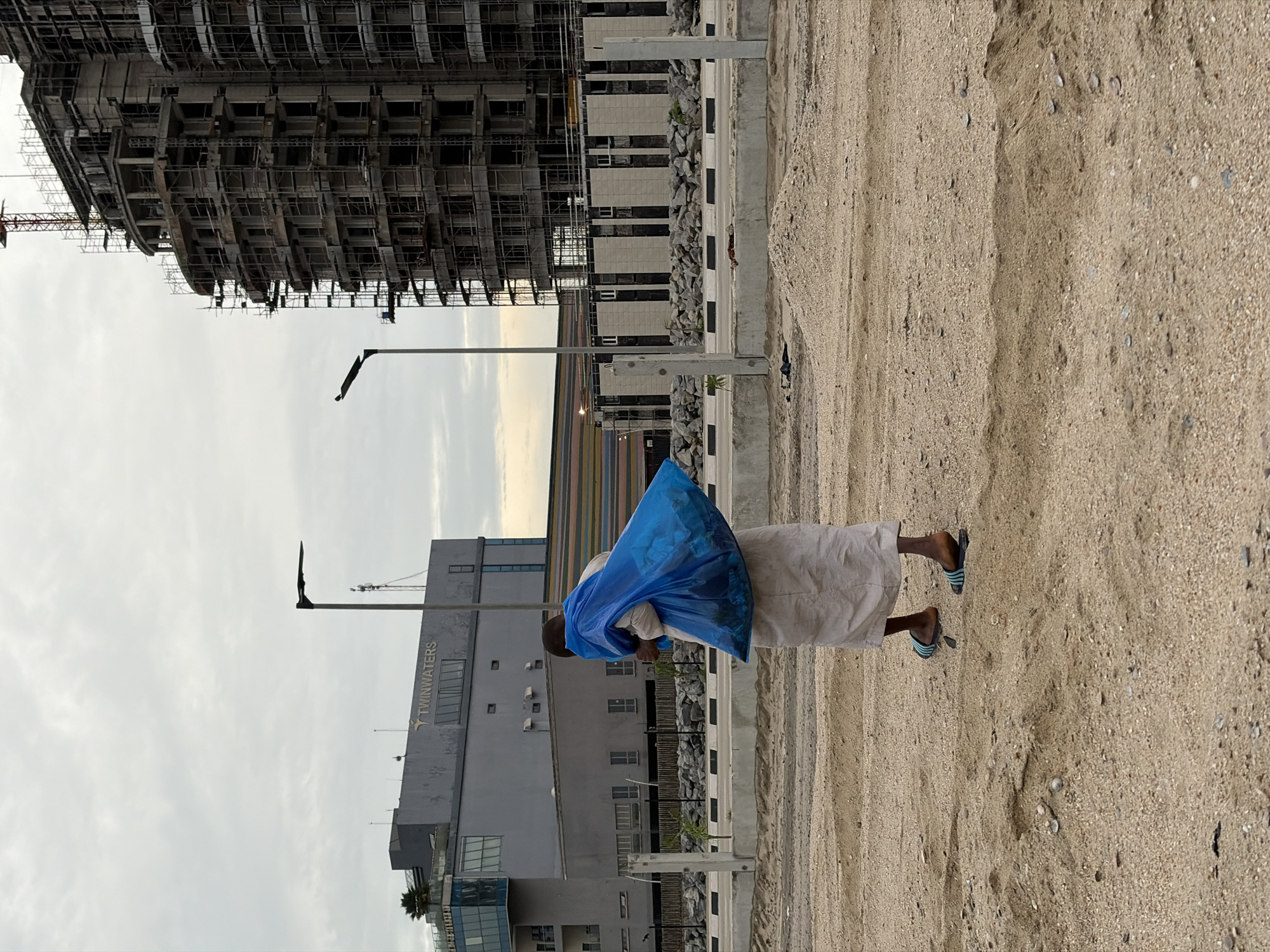 A full-body, rear-view shot captures an individual walking across a sandy, undeveloped area, carrying a large, bright blue plastic bag over their left shoulder. The person is wearing a long, light-colored garment that extends to their ankles and what appear to be flip-flops on their feet. The background features a stark contrast between ongoing construction and completed buildings. To the left, a multi-story building with the signage "TWINWATERS" is visible, with a palm tree on its roof. Adjacent to this, another grey building with numerous windows is seen. Dominating the right side of the frame is a large, unfinished building, heavily wrapped in scaffolding, with a red crane extending from its top. The ground is predominantly composed of sand and small pebbles, with some scattered rocks and concrete barriers in the middle ground. The sky is overcast with a pale, diffused light. This scene is located in Alaguntan, Nigeria, a developing urban area characterized by construction and a mix of completed and in-progress structures.