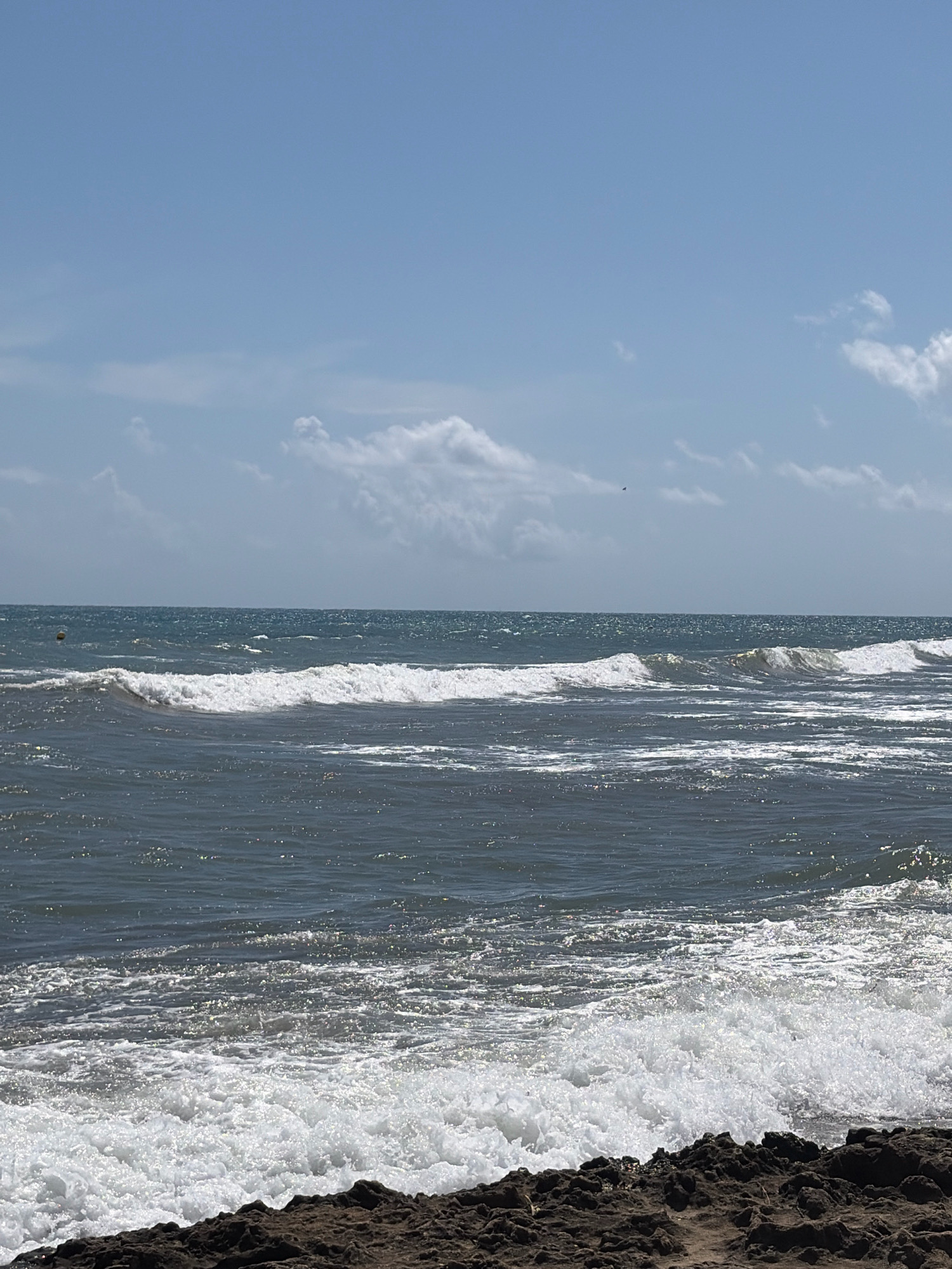 A bright, sunny day at the beach in Torrevieja, Spain. The image shows a vast expanse of the Mediterranean Sea, with waves crashing against a rocky shore in the foreground. The water is a deep blue, with white foam from the waves creating a stark contrast. The sky is a clear blue with scattered white clouds. There are no people visible in the image. The rocky shore is covered in dark, textured rocks, with some sand visible between them. The scene is tranquil and captures the natural beauty of the coastline.